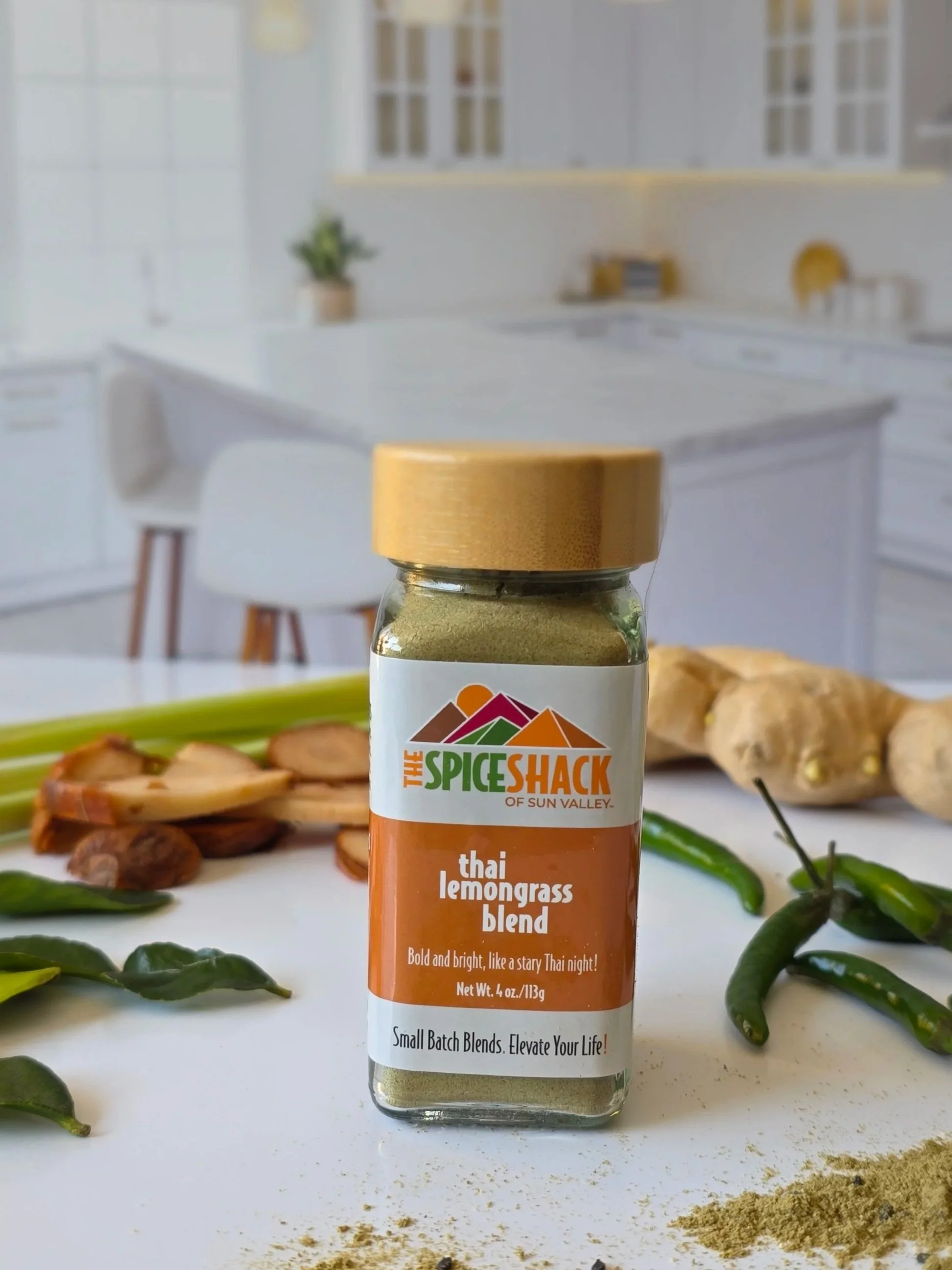 Bottle of Thai lemongrass blend spice mix surrounded by lime slices, lemongrass stalks, green chili peppers, ginger pieces, black lime leaves, and turmeric powder on a white surface.