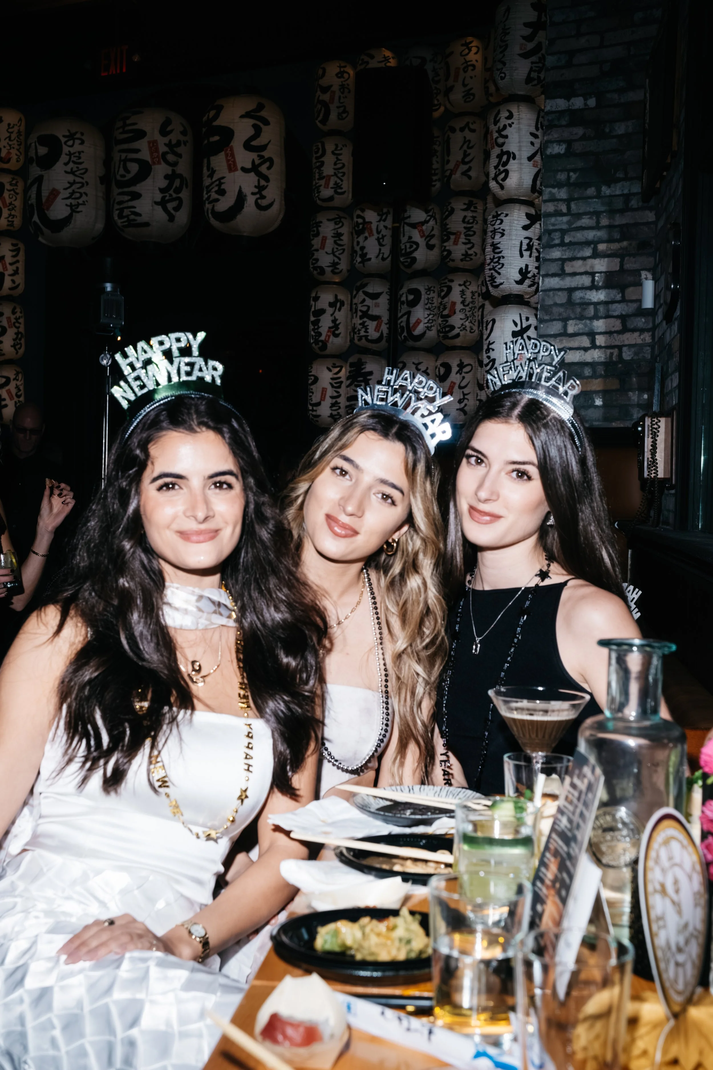 Three women celebrating New Year's Eve at a restaurant, wearing 'Happy New Year' headbands, sitting at a table with food and drinks, with lanterns and Japanese writing in the background.