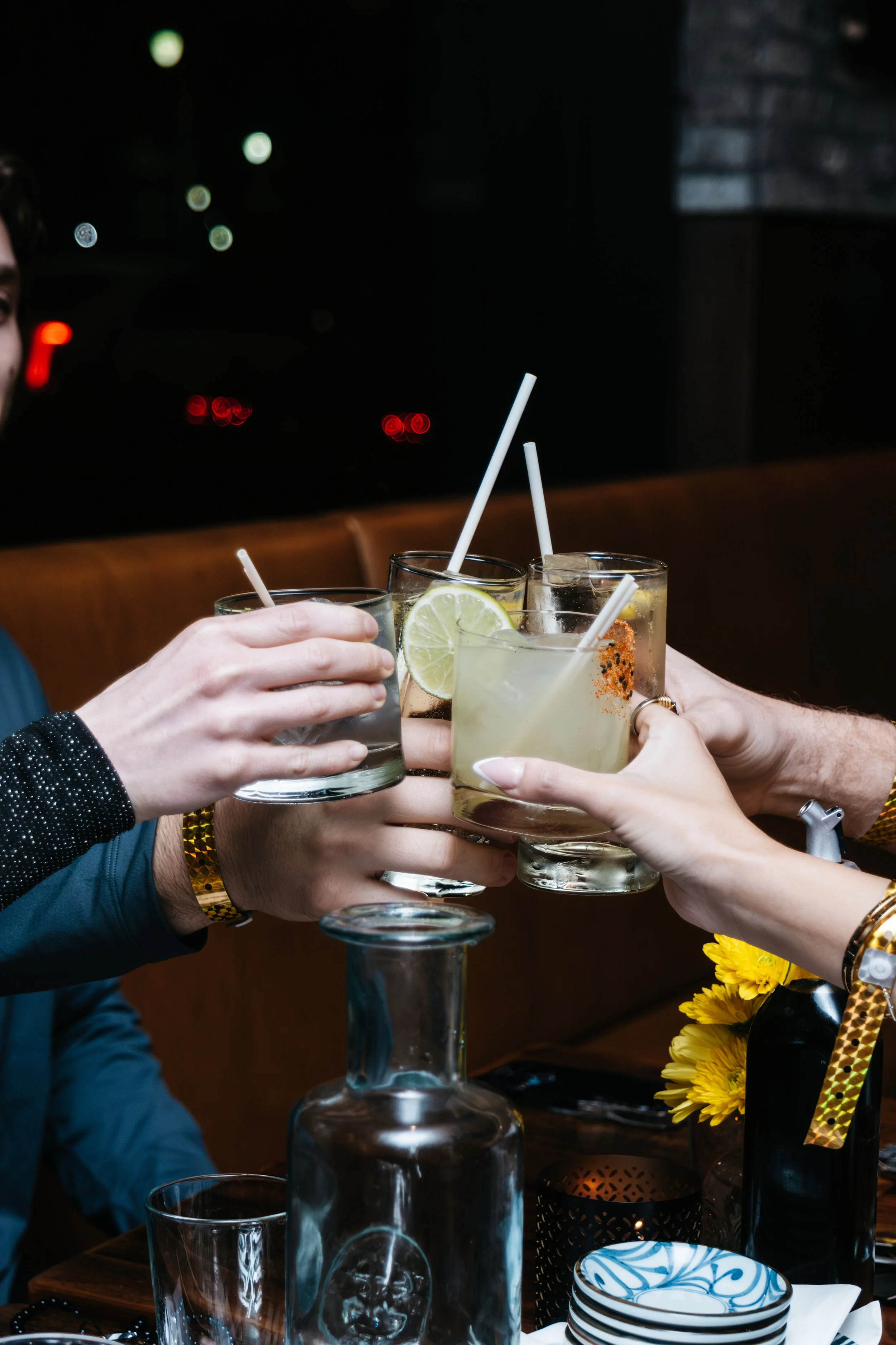 Group of people clinking glasses with cocktails, some garnished with lemon slices, in a dimly lit restaurant or bar setting.