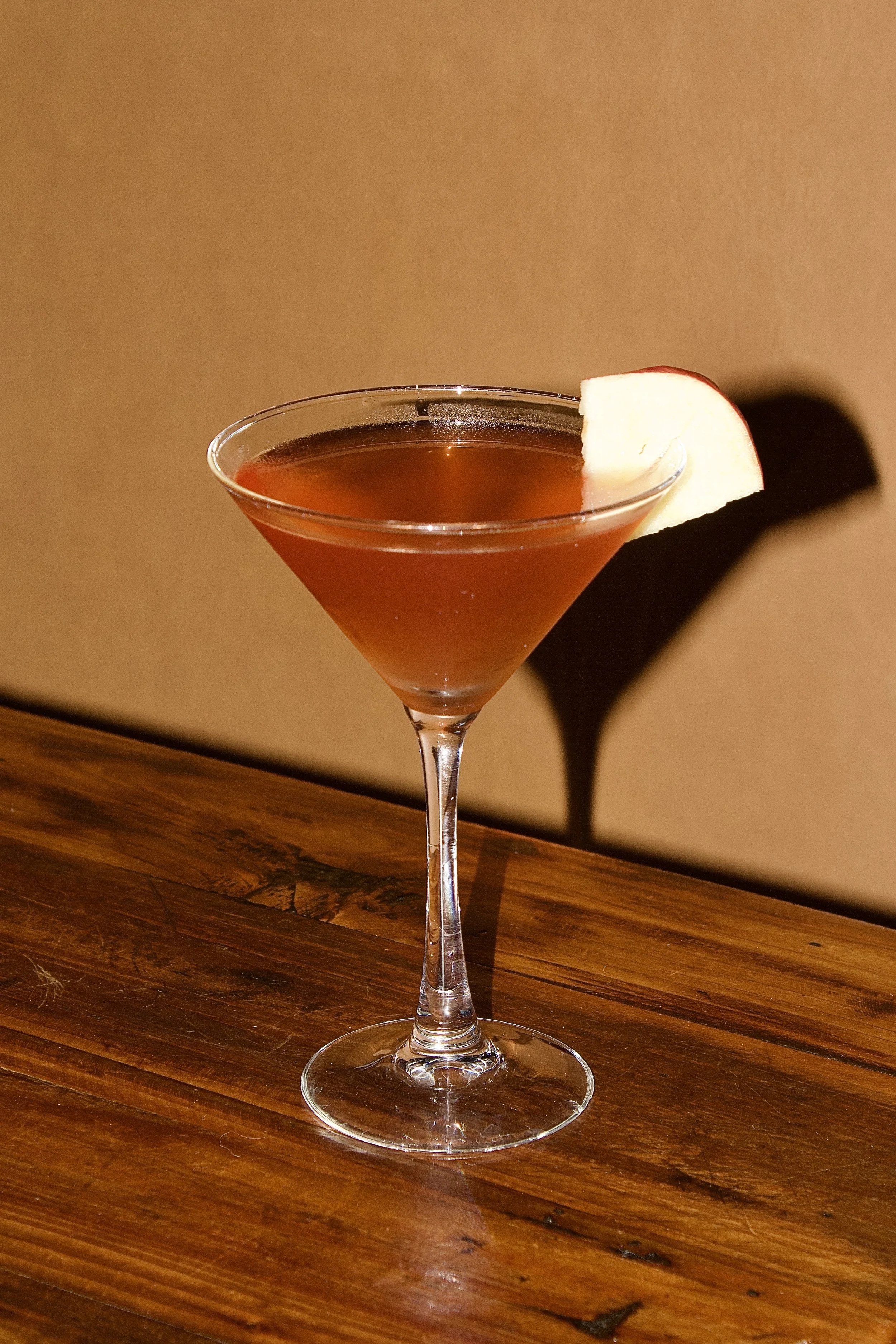 A cocktail in a martini glass with a slice of apple as garnish, placed on a wooden surface with a plain background.