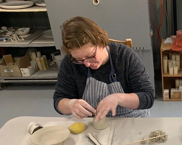 a woman wearing an apron is sitting at a table  scupting clay with her hands.
