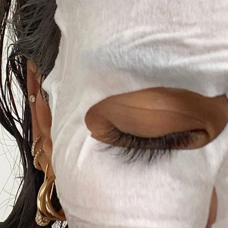 Close-up of a person with dark, wavy hair and multiple earrings, covered with a white facial mask, with eyes closed.