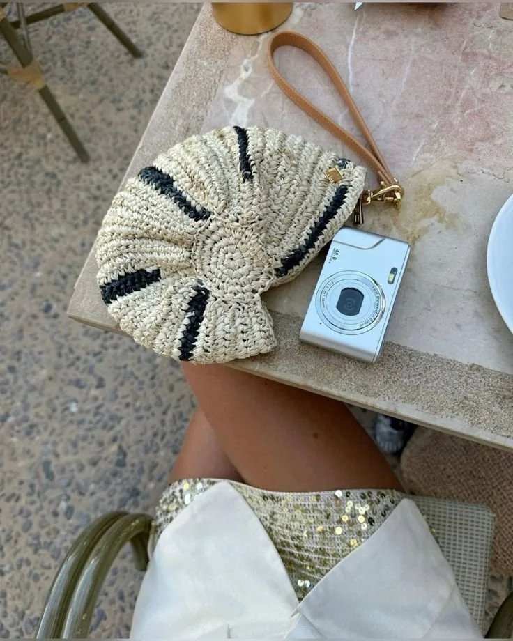 A beige woven purse with black stripes, a leather wristlet strap, and a small metal charm, resting on a table. Next to it is a small silver digital camera. The person's lap is visible, wearing a white dress with a gold sequined trim. The table has a plate on one side and a brass object in the background. The setting appears to be outdoors or in a casual dining area.