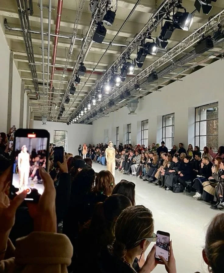 A fashion runway show with seated audience on both sides, a model walking on the runway, and photographers capturing the runway from the audience.