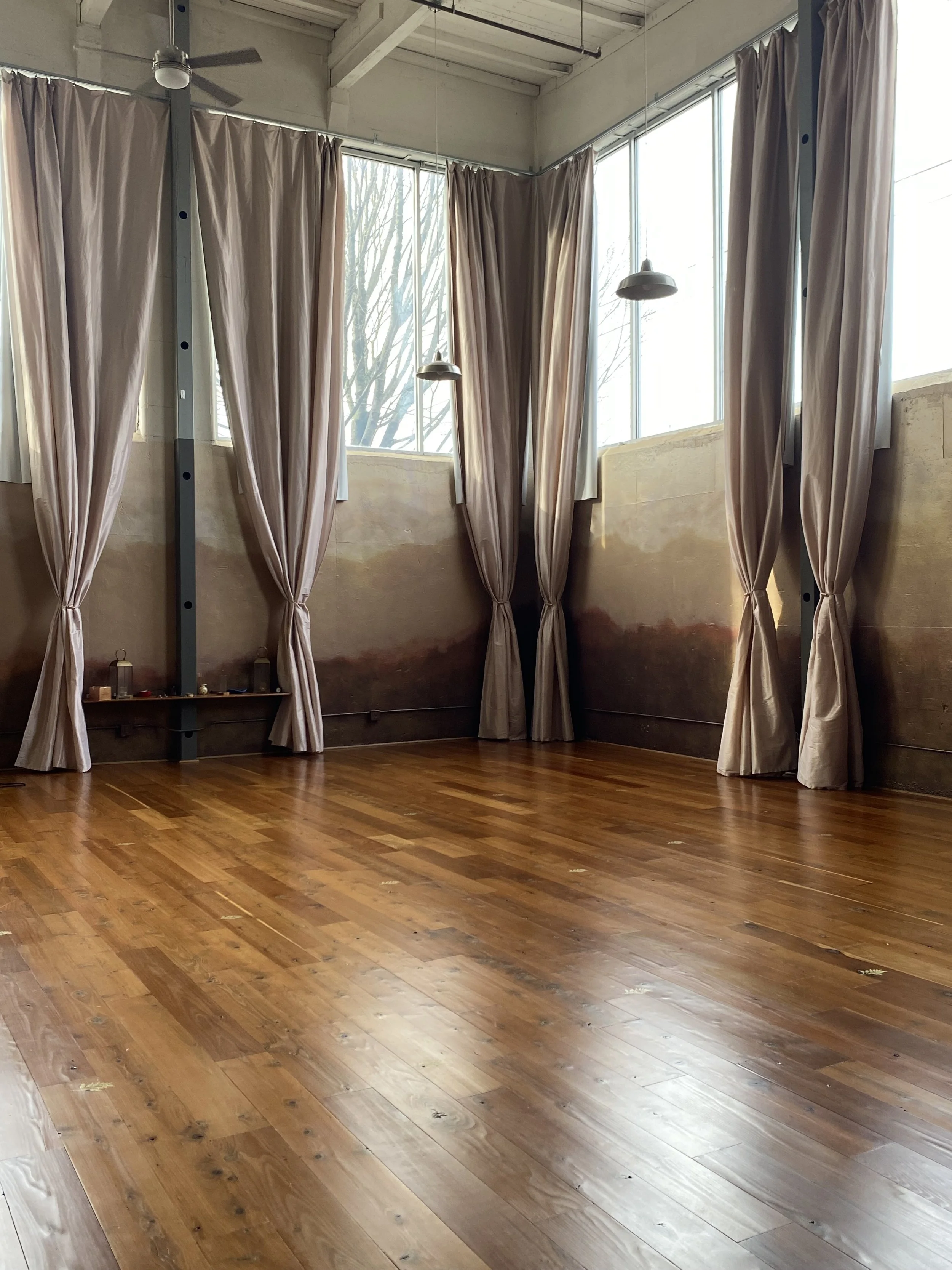 Empty room with large windows, beige curtains, and wooden flooring.