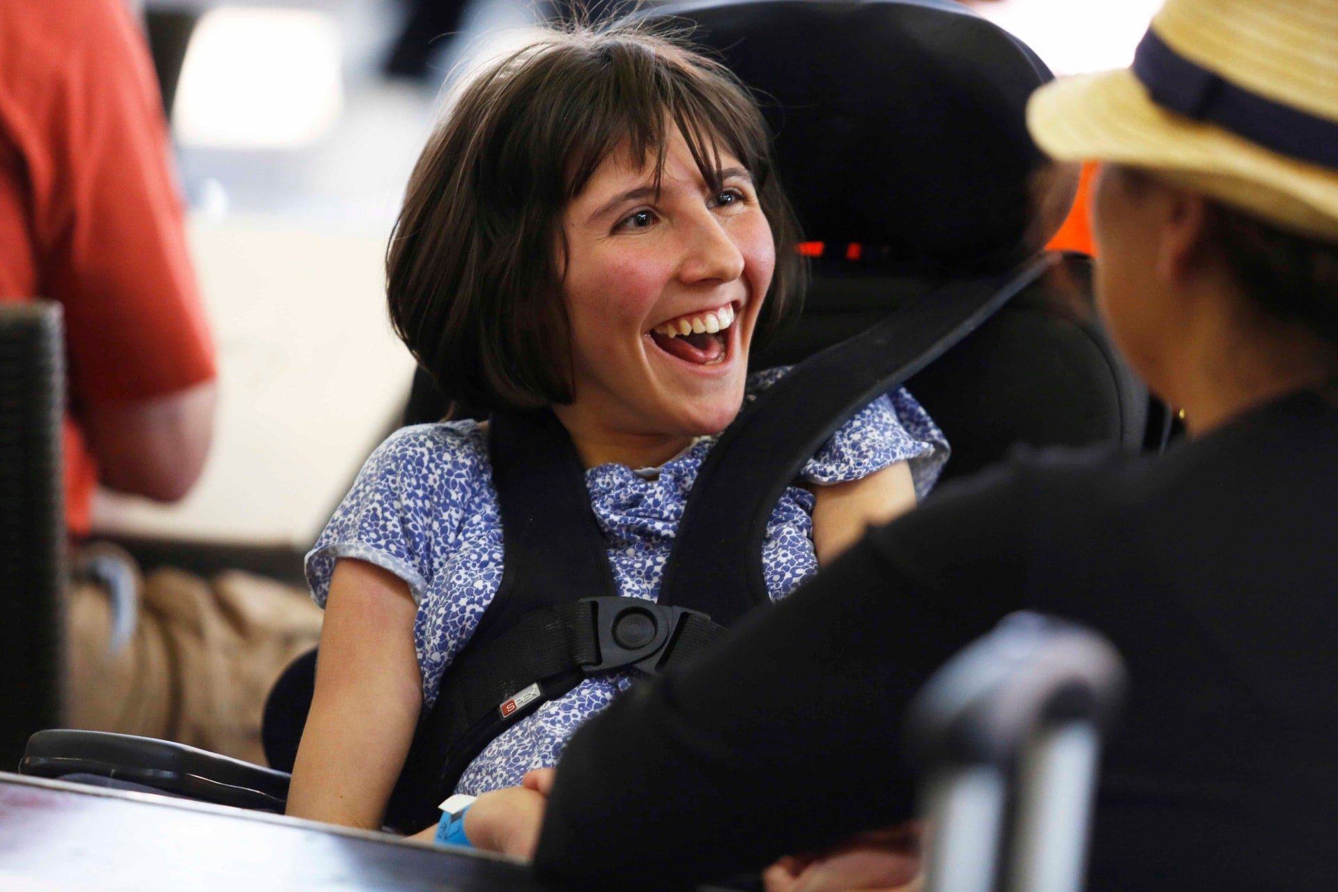 A woman in a blue patterned outfit in a wheelchair smiling and looking at another person wearing a straw hat.