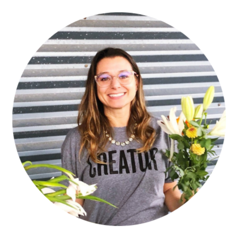 A woman with brown hair, glasses, and a grey shirt that says 'CREATOR' holding a bouquet of white lilies and yellow flowers, standing in front of a metallic striped background.