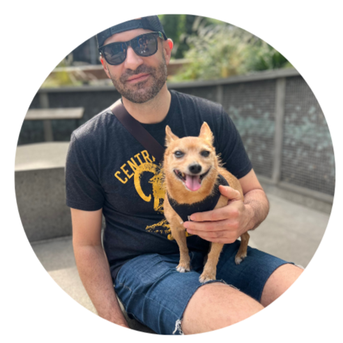 A man wearing sunglasses, a black t-shirt, and a cap, sitting outdoors, holding a small tan dog with a happy expression.
