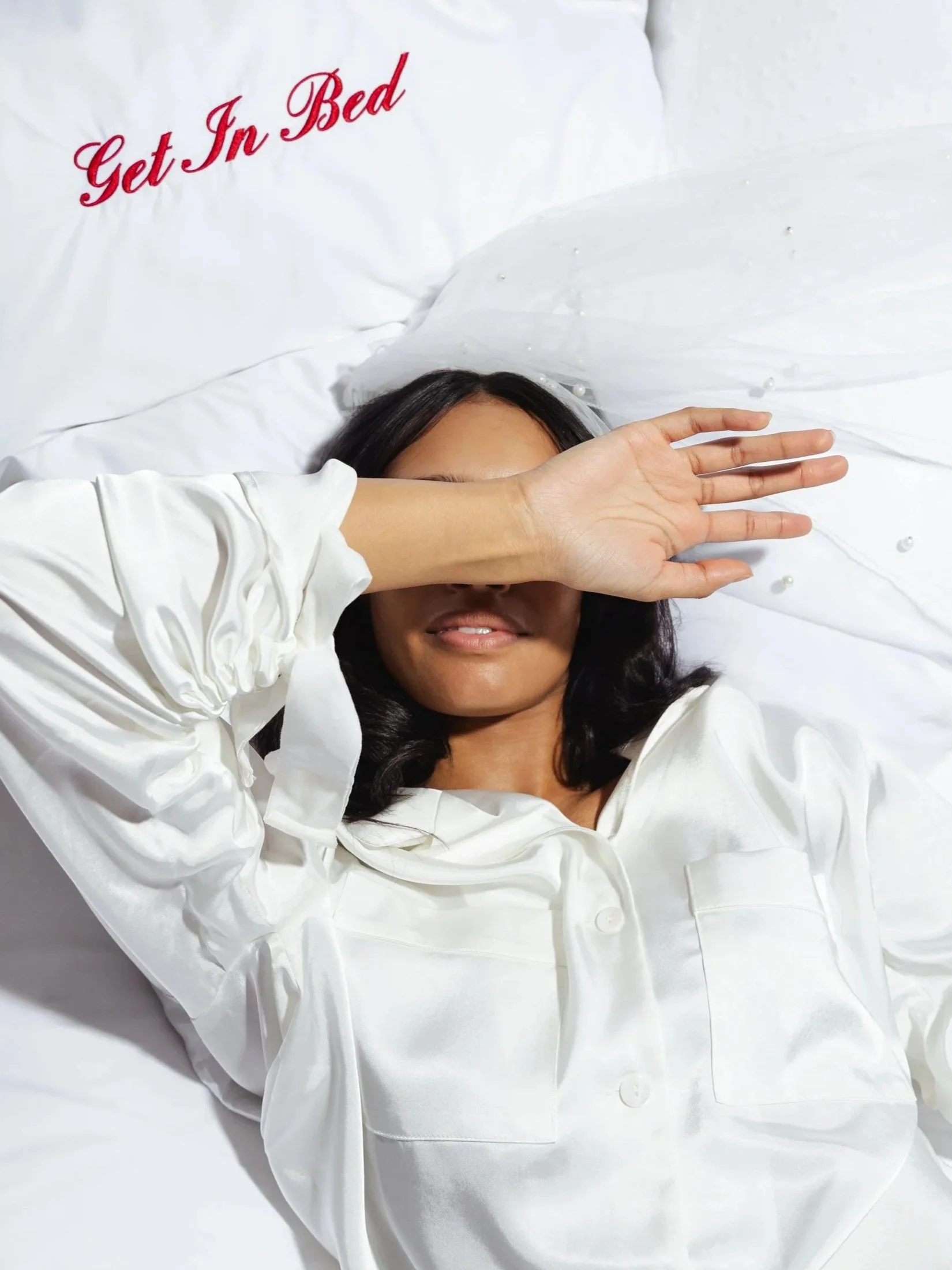 Woman lying in bed with a white satin pillow and sheet, partially covering her face with her arm. The pillow has red embroidered text that reads 'Get In Bed.' She has dark hair, smiling, and is wearing a white satin or silk robe.