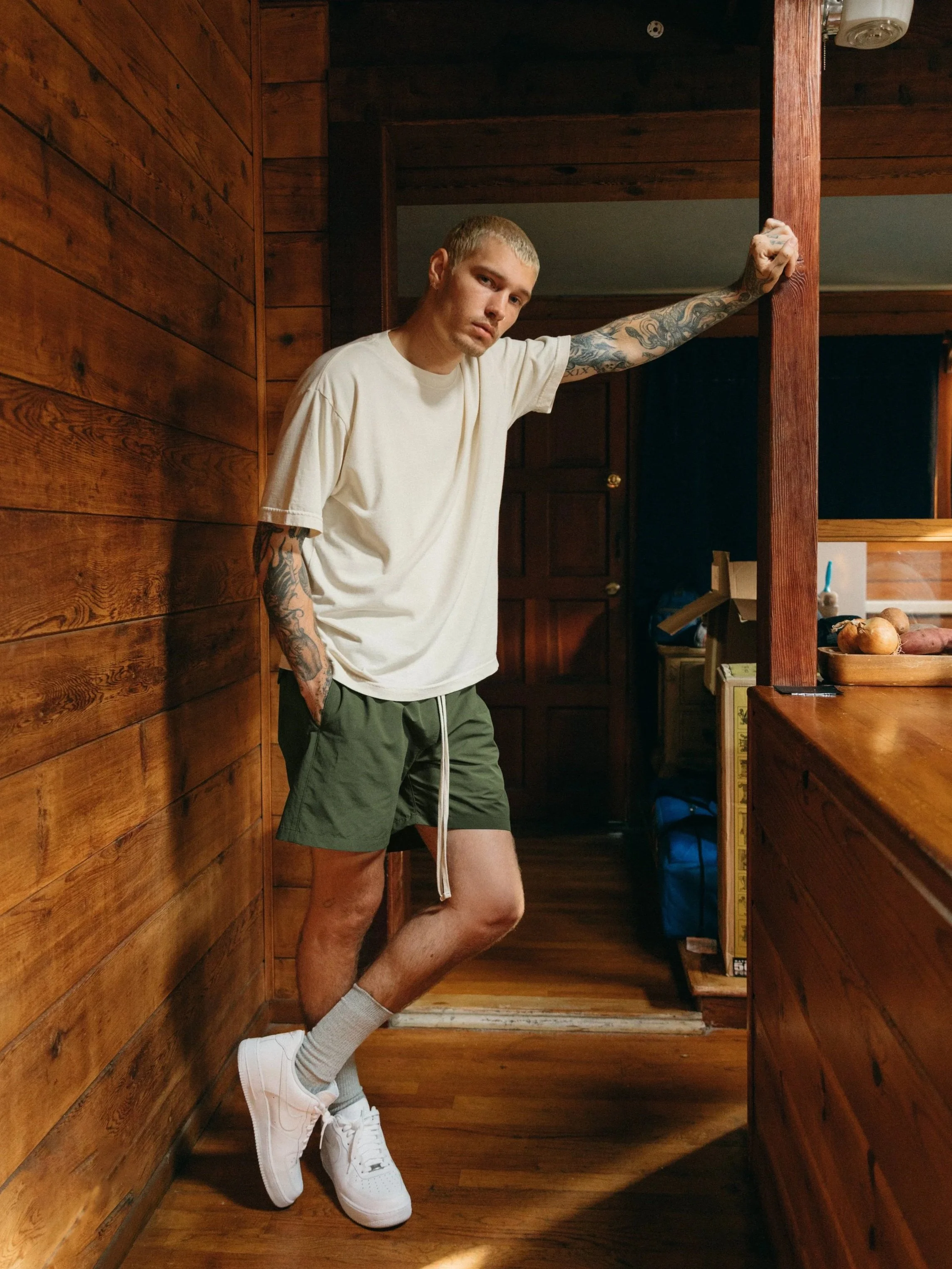 Young man with tattoos wearing a white t-shirt, green shorts, white sneakers, and beige socks, standing in a wooden kitchen, leaning against a wooden beam with a serious expression.