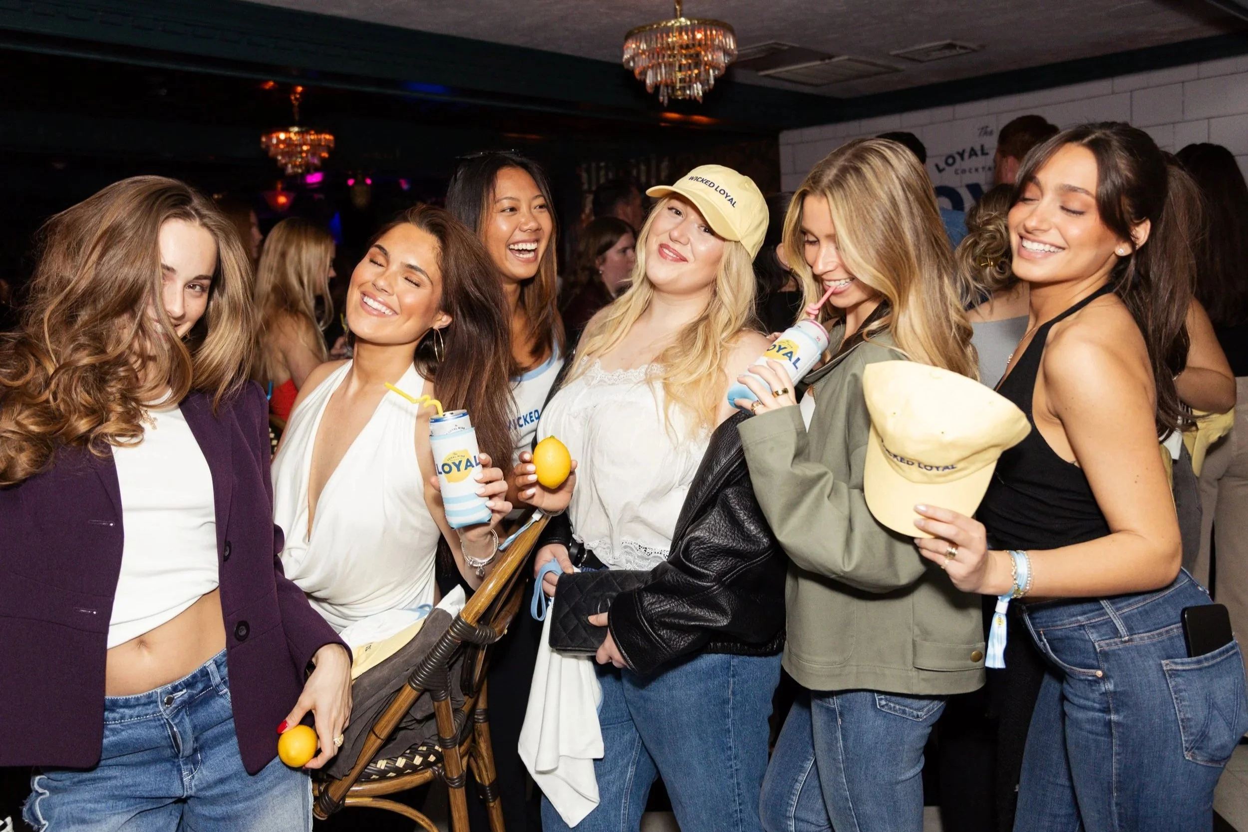 Group of women at a social event, smiling and standing close together, some holding beverages and wearing casual clothing.