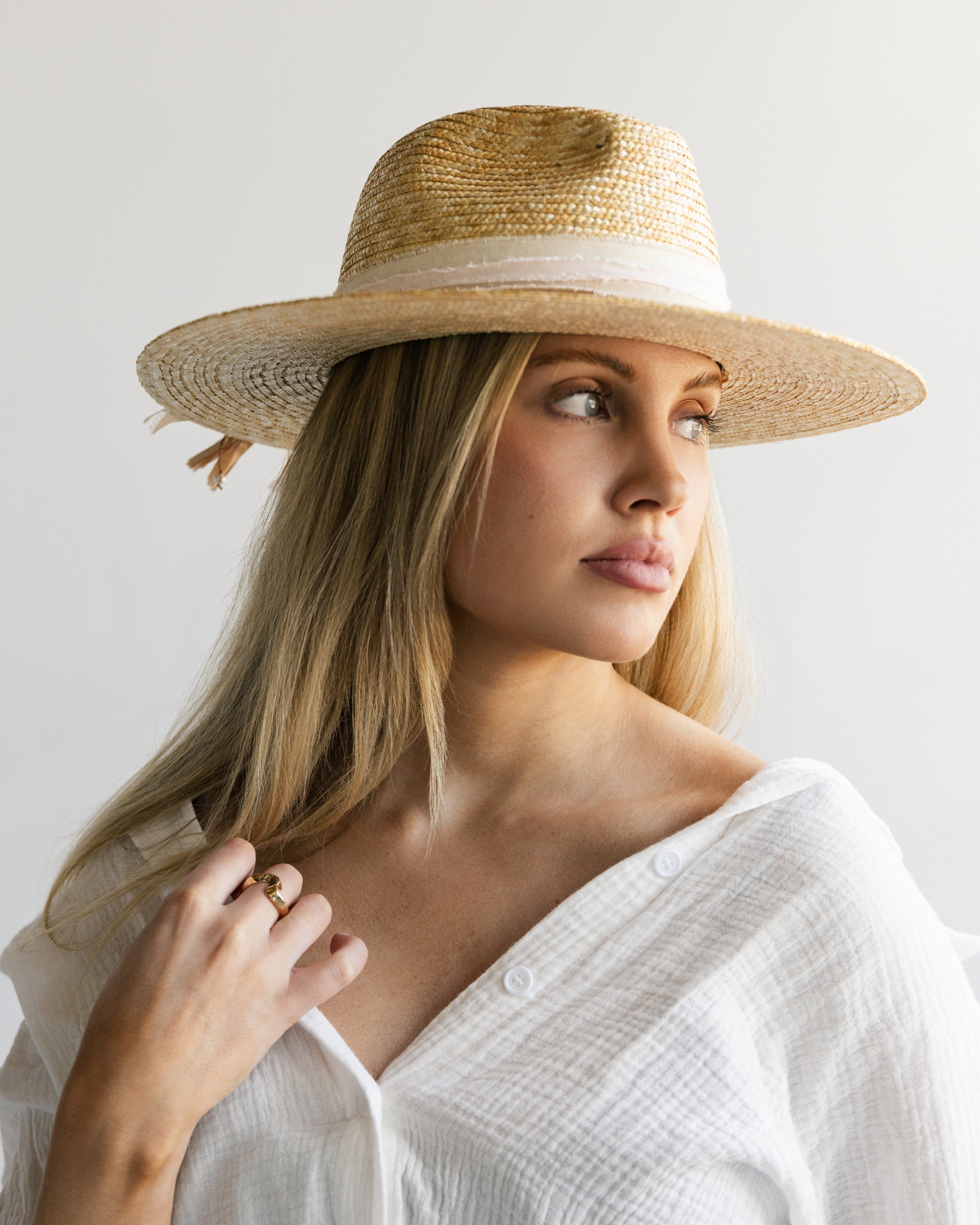 A young woman with blonde hair wearing a wide-brimmed straw hat and a white button-up linen shirt, standing against a plain, light background.