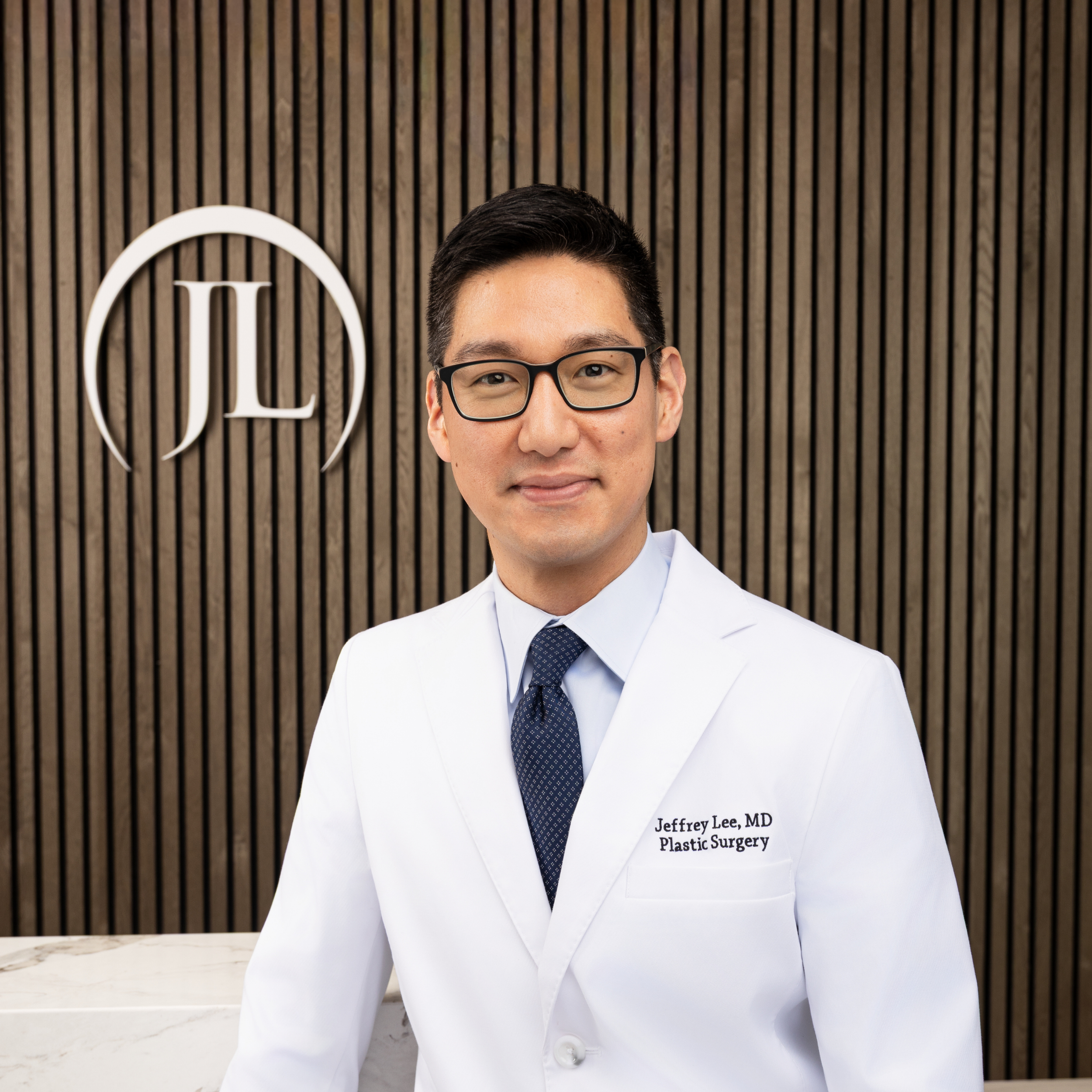 Close-up of a male doctor wearing glasses and a white coat with 'Jeffrey Lee, MD Plastic Surgery' embroidered on it, standing in front of a vertical wooden slat wall with a white circular logo.