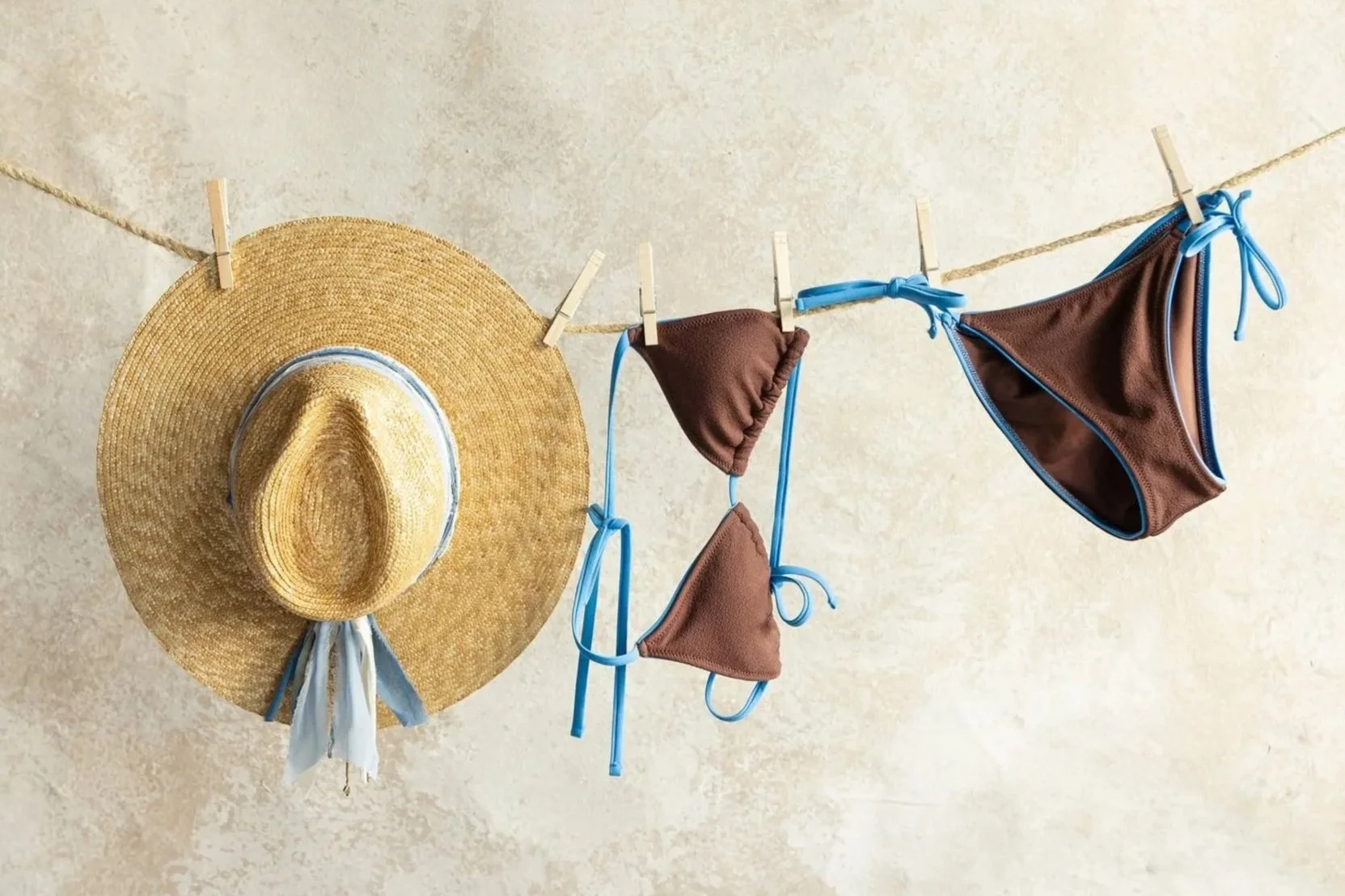 A straw sun hat and a brown bikini with blue trim hanging on a clothesline against a beige wall.