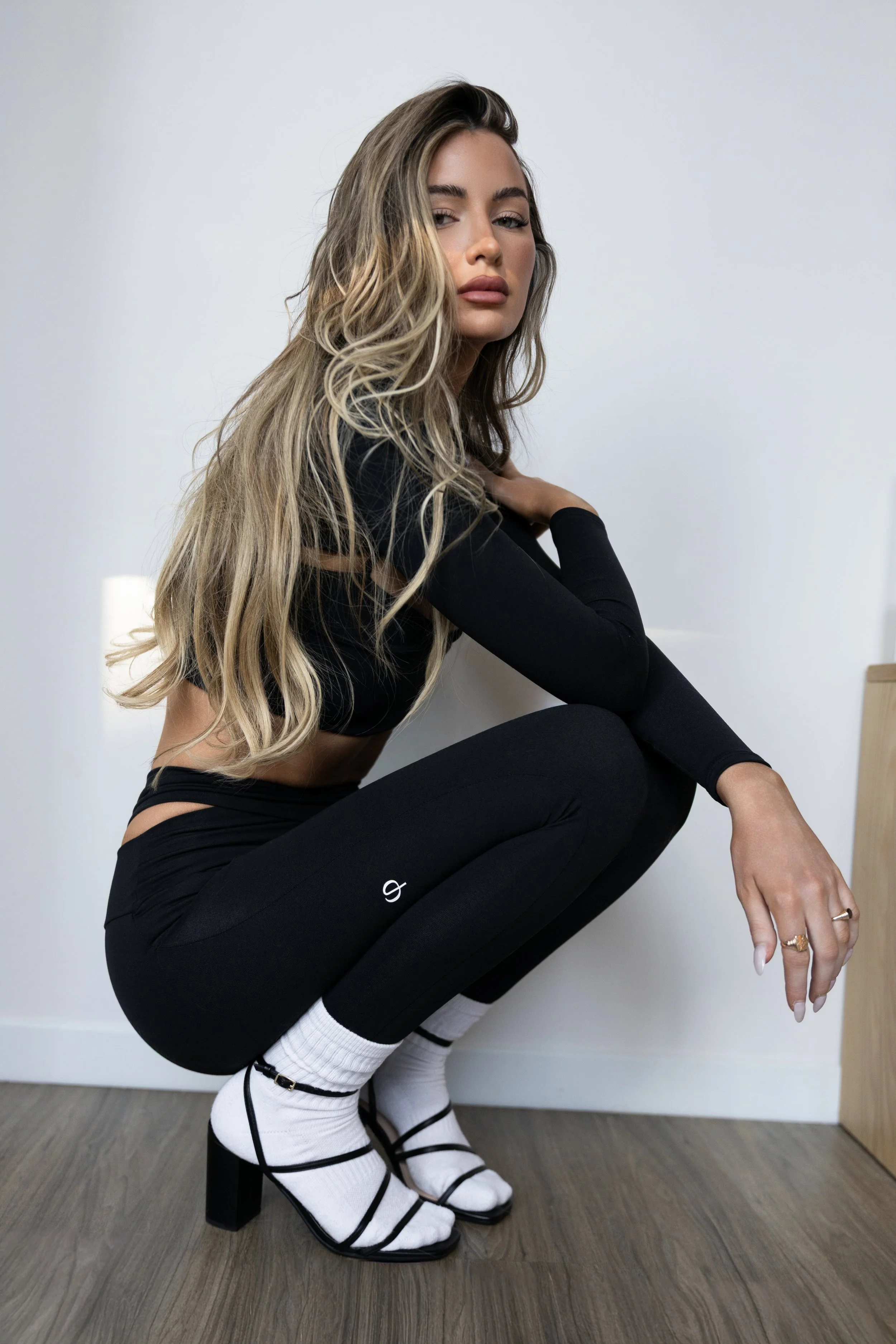 A woman with long, wavy blonde hair squatting on the floor, wearing black leggings, white knee-high socks with black stripes, strappy black heel sandals, and a black long-sleeve top, positioned against a plain white wall.