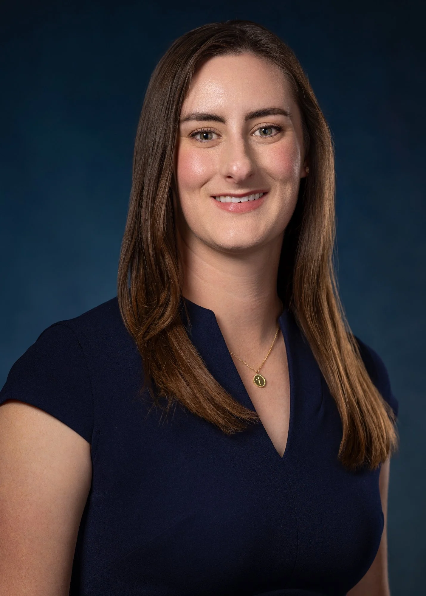 Professional head-and-shoulders portrait of a smiling woman with long brown hair wearing a navy dress and gold pendant necklace against a dark blue background