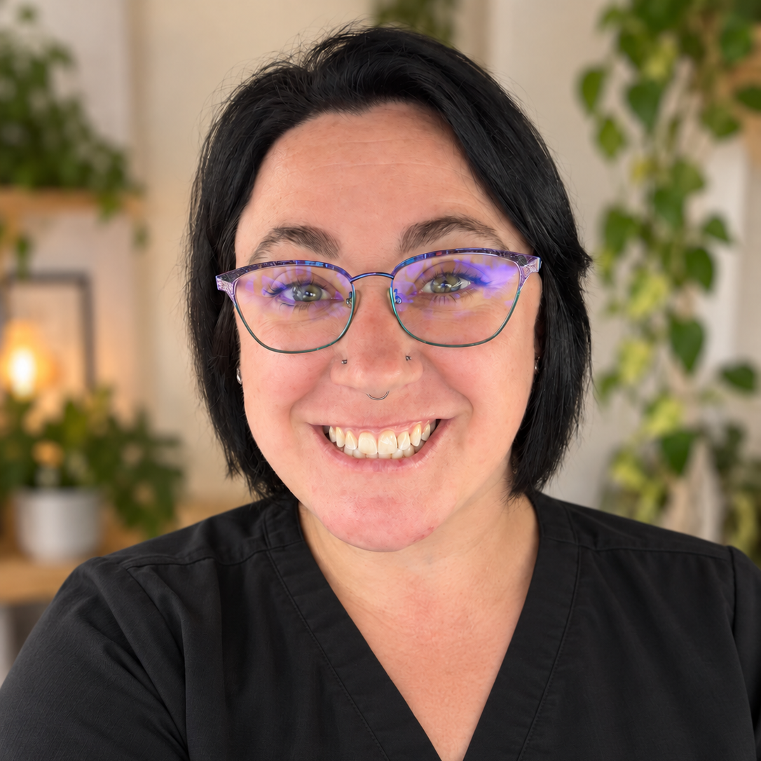 A smiling woman with short black hair, wearing glasses with purple frames, and a black top, indoors with green plants in the background.
