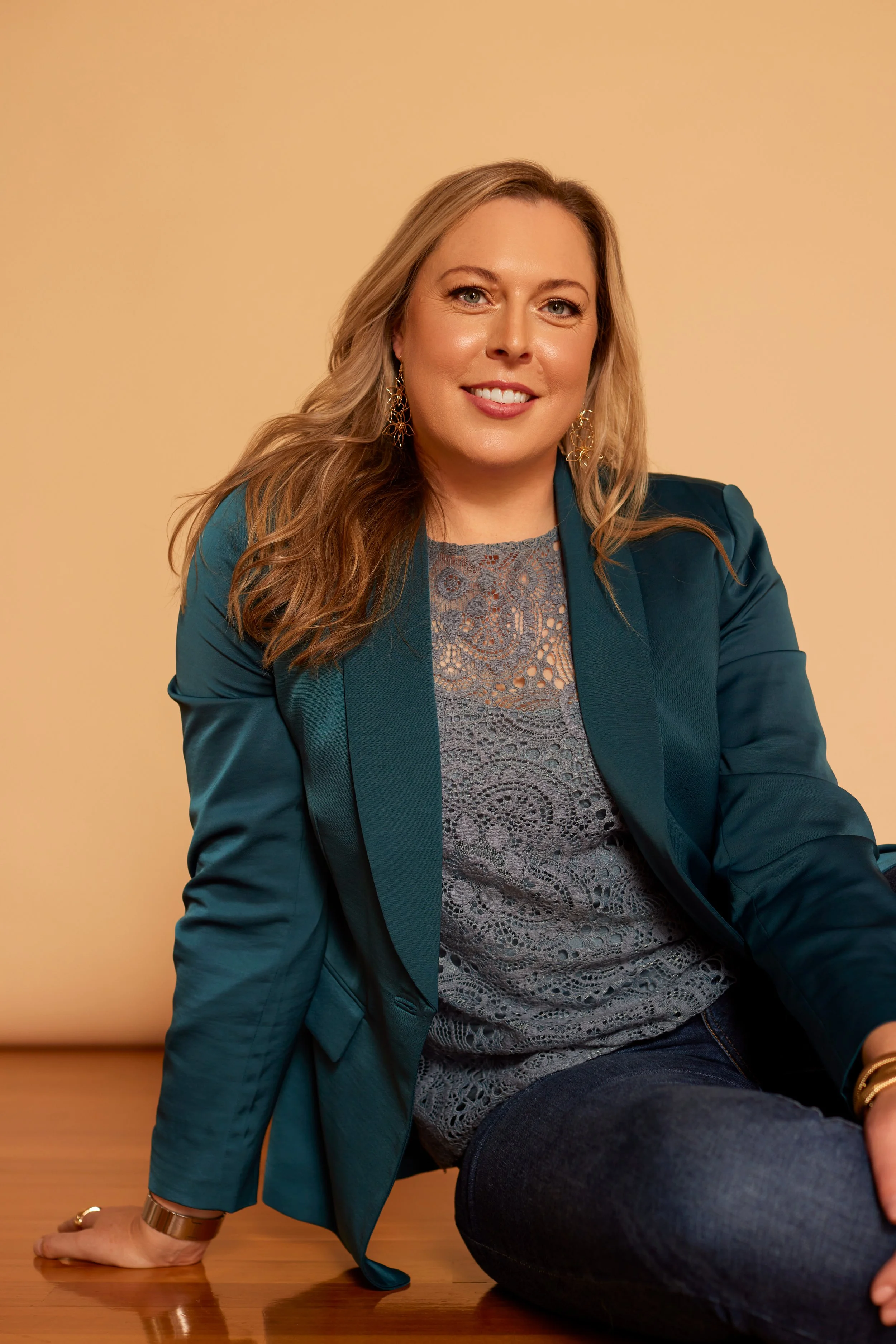 A woman with long, wavy blonde hair sitting on a wooden floor, wearing a teal blazer, gray lace top, dark jeans, and gold jewelry, smiling at the camera against a beige background.