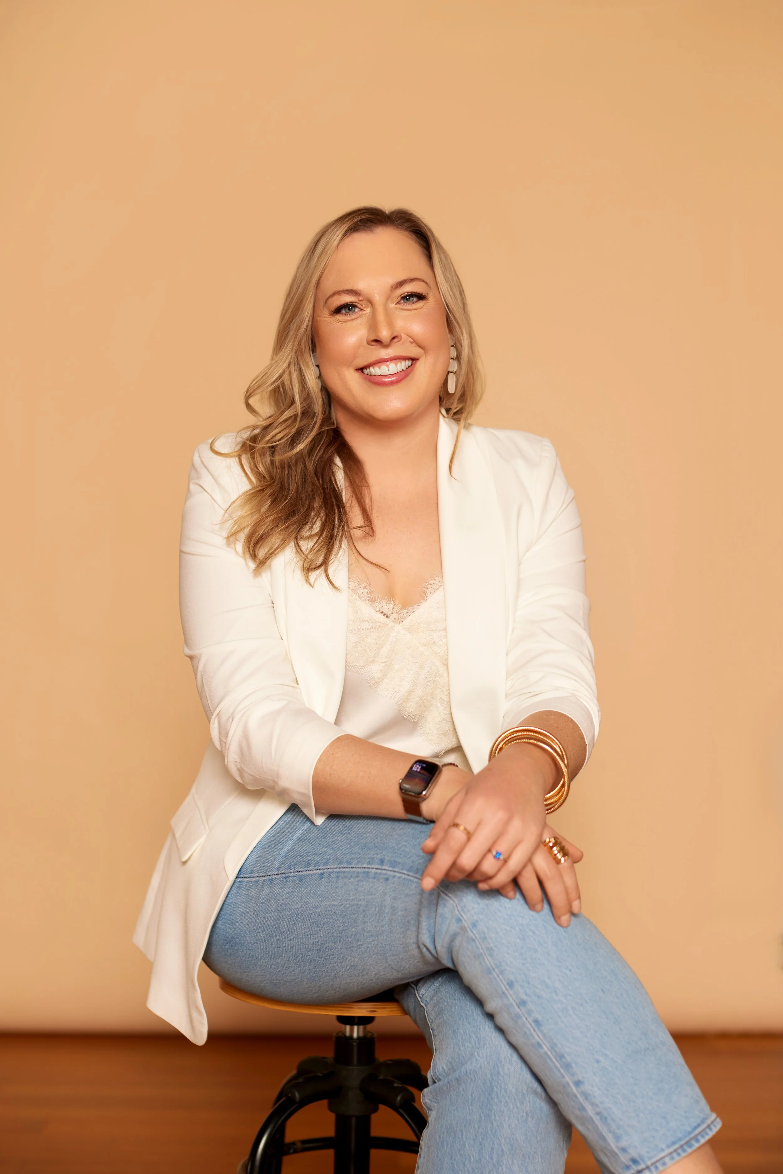A woman with blonde hair sitting on a stool against a beige background, smiling at the camera, wearing a white blazer, lace top, blue jeans, and accessorized with bracelets, rings, earrings, and a smartwatch.