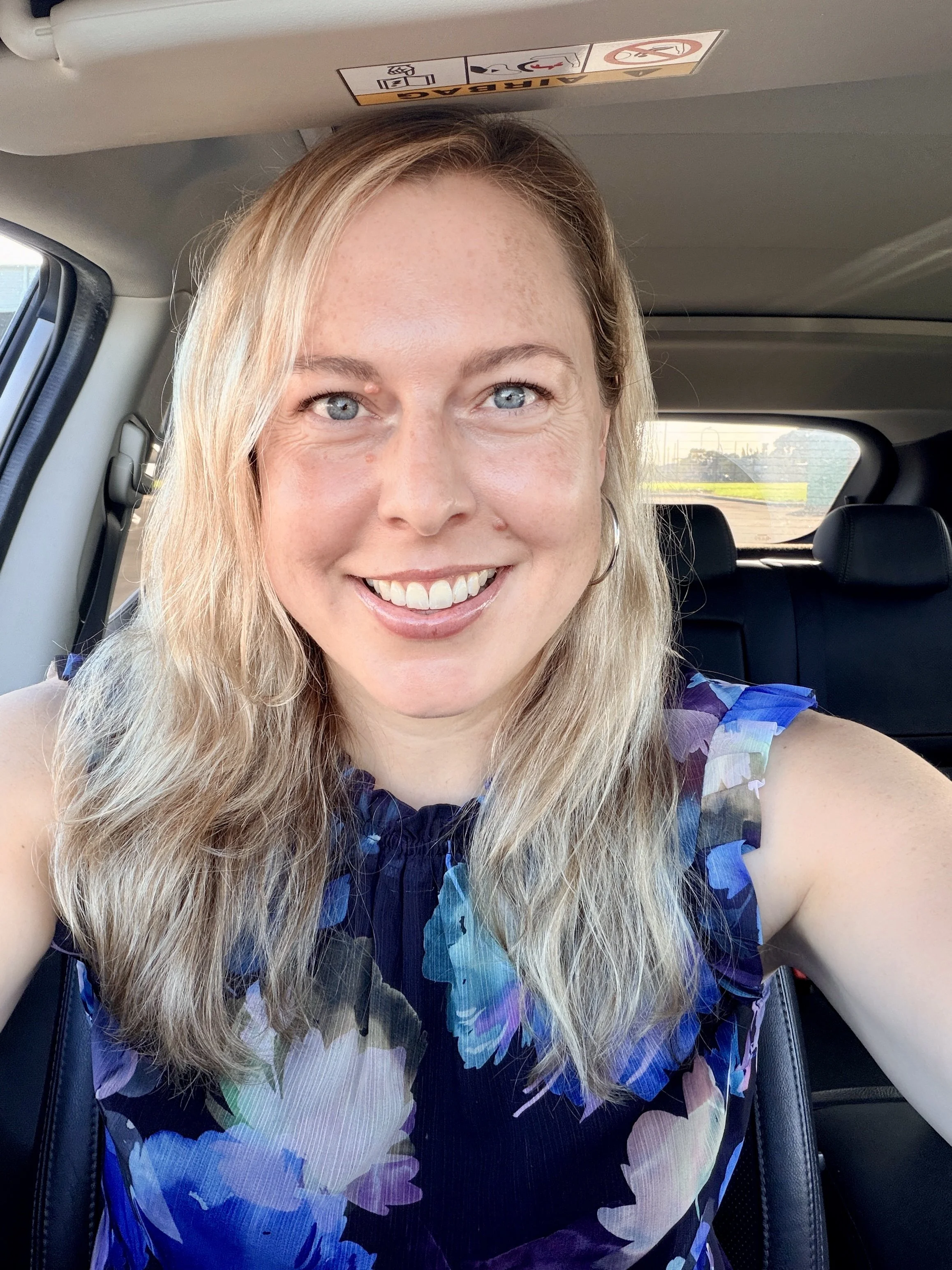 Close-up of a woman with blonde hair and blue eyes, smiling, sitting in a car with a patterned blue and purple top.