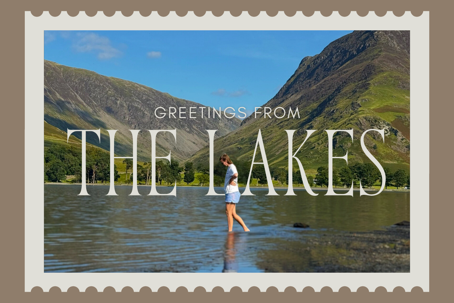 Large lake at the base of two large green mountains. A woman in a white t-shirt and blue shorts walks through he water. Behind her, white lettering spells out "GREETINGS FROM THE LAKES"