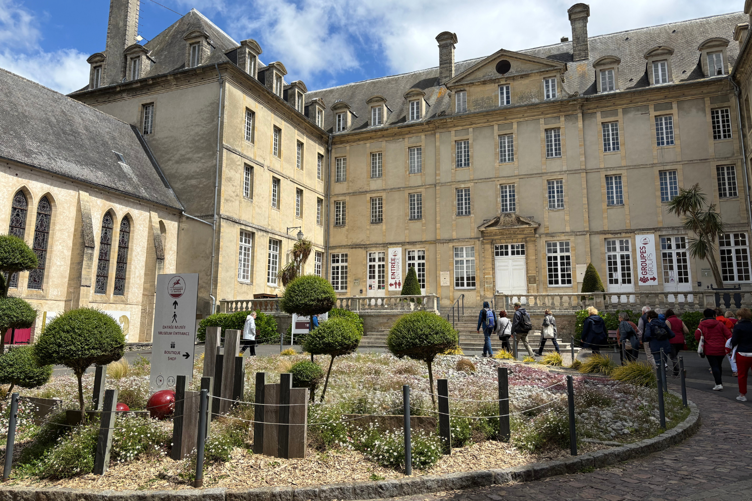 Large tan building with three rows of several windows. A circle of wildflowers sits in the middle of a pathway that leads to the entrance of the building.
