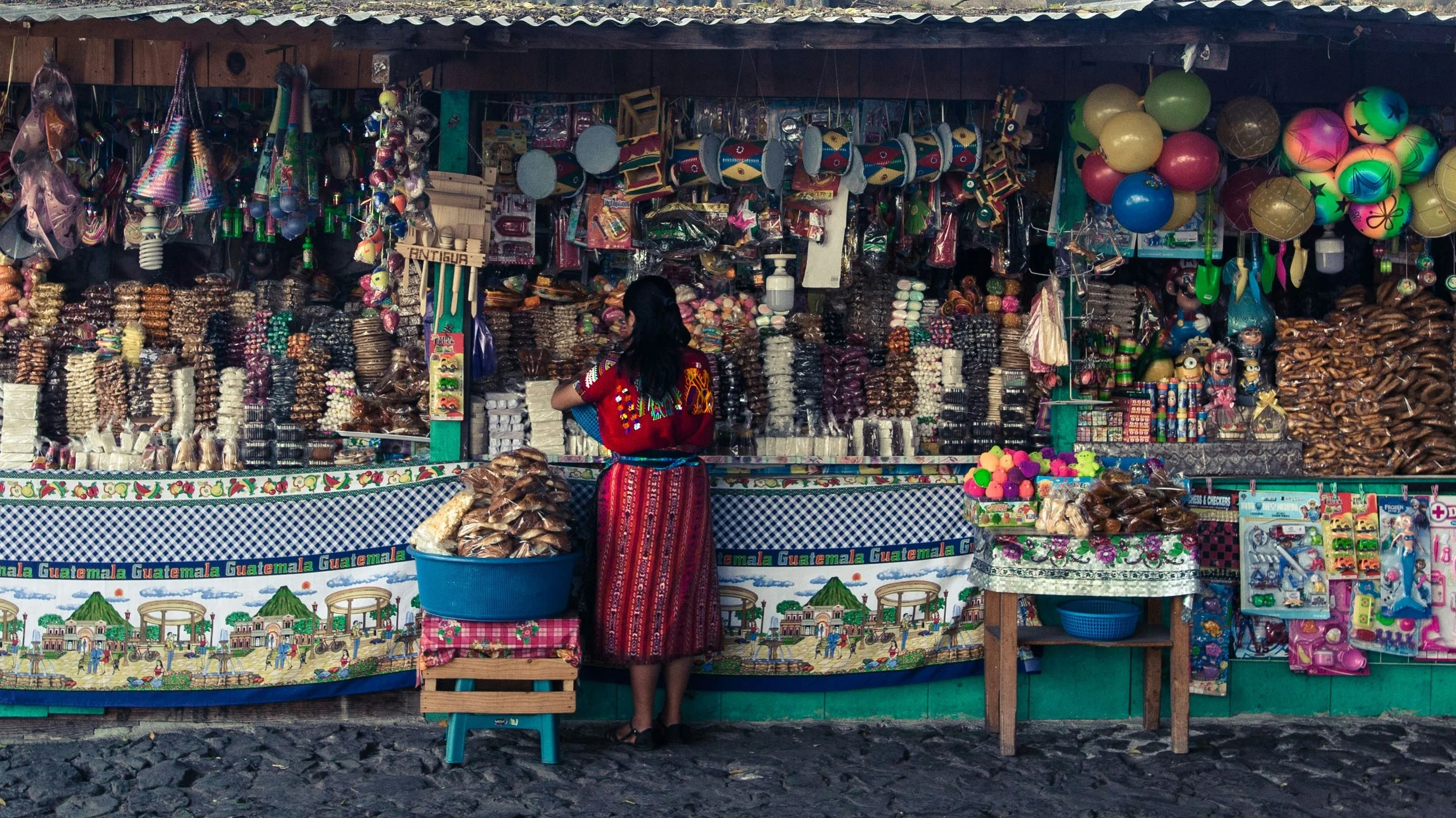 Woman in colorful red dress looks over a food stall that has stacks upon stacks of bags of beans, candied nuts, etc. There is a table cloth visible that reads the word "GUATEMALA: over and over again