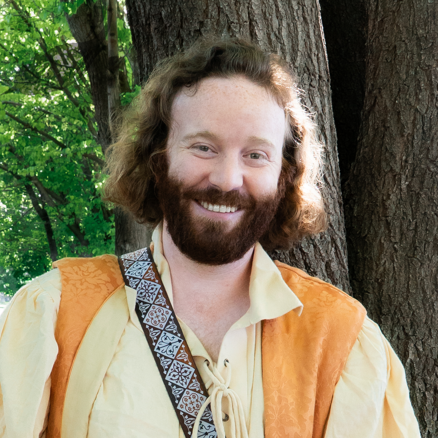 A man with long, curly hair and a beard, smiling, standing outdoors in front of a tree with green leaves.