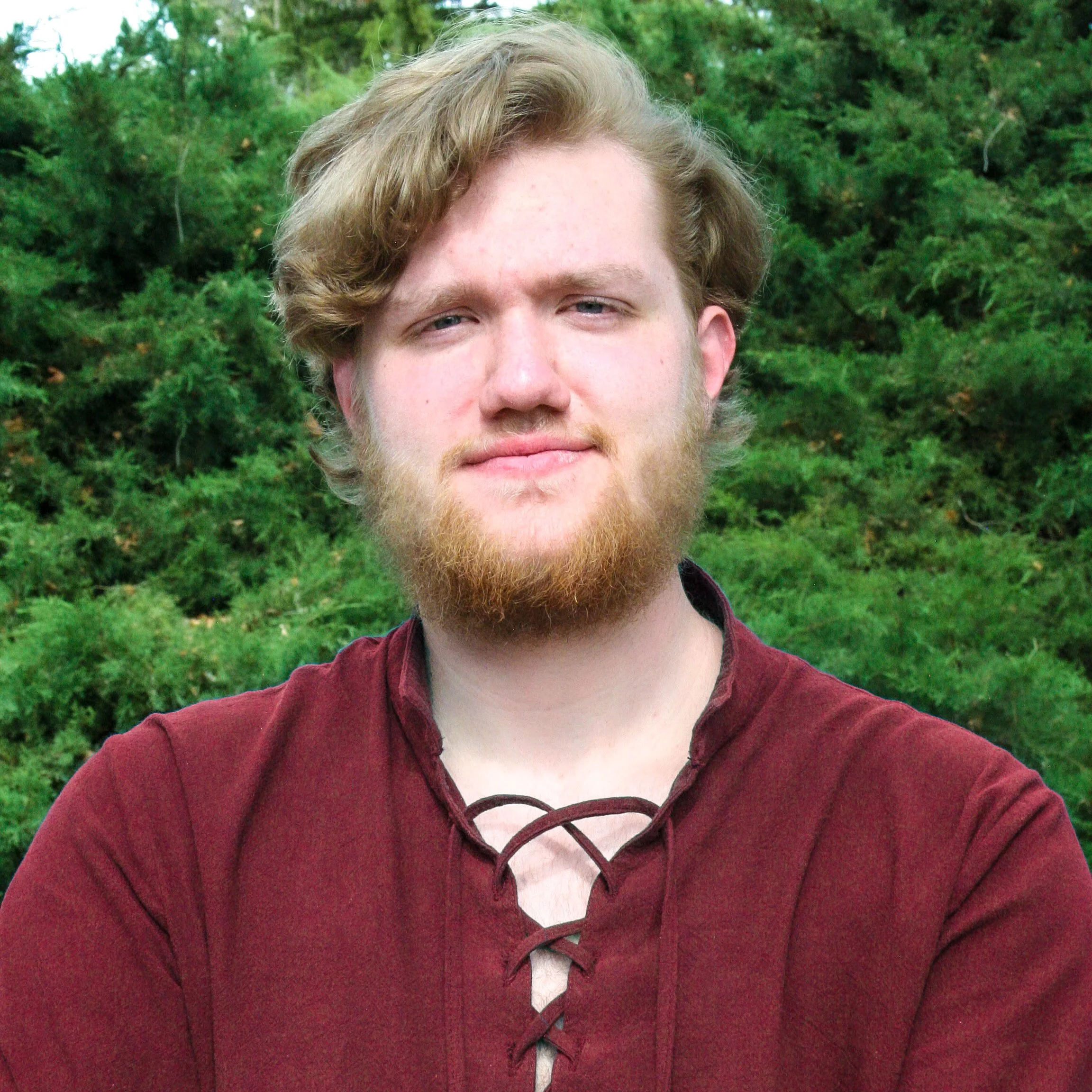 Close-up of a young man with blonde hair and a beard wearing a maroon shirt, outdoors with green trees in the background.