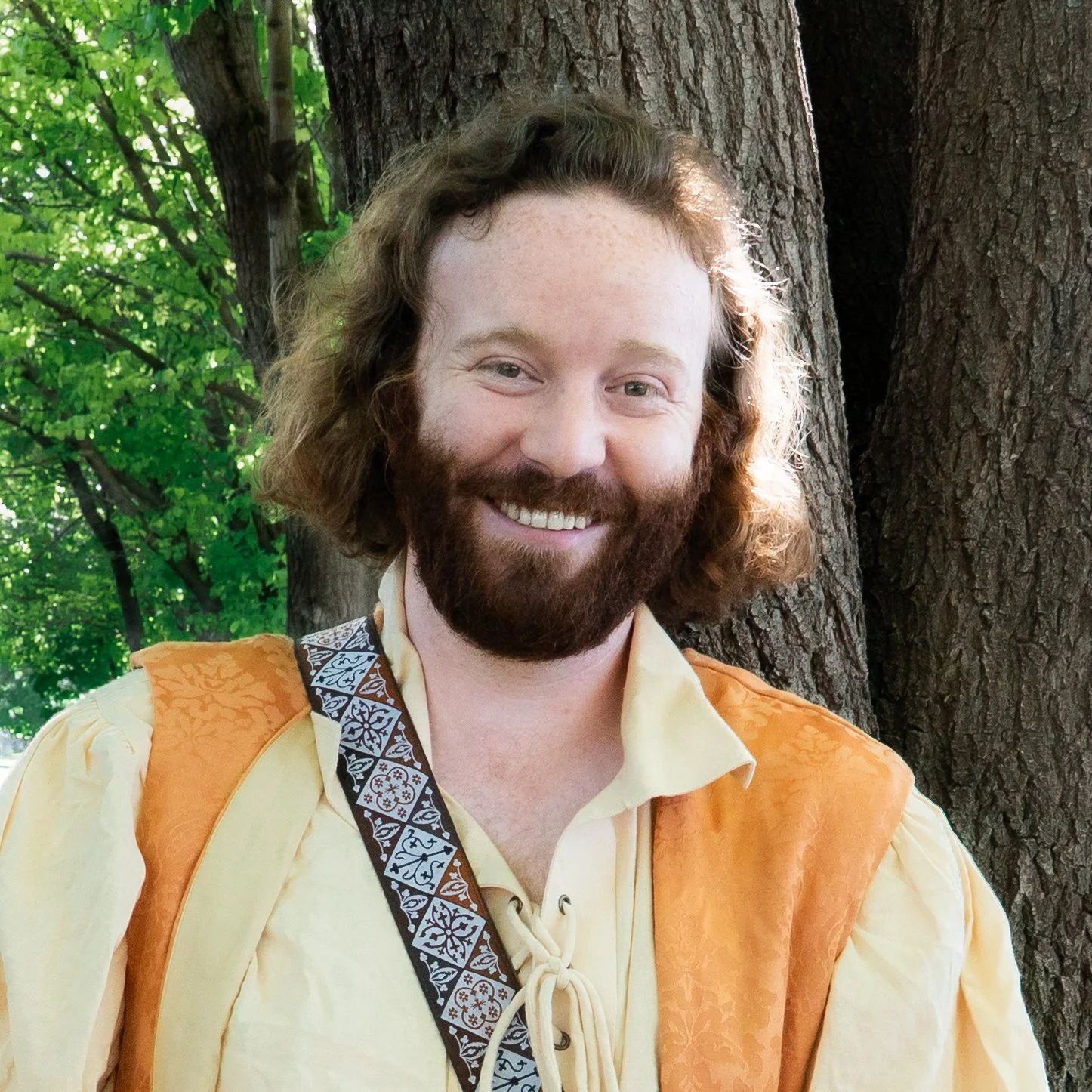 A smiling man with a beard and long hair standing outdoors in front of a tree, wearing a yellow shirt and an orange vest, with a strap across his chest.