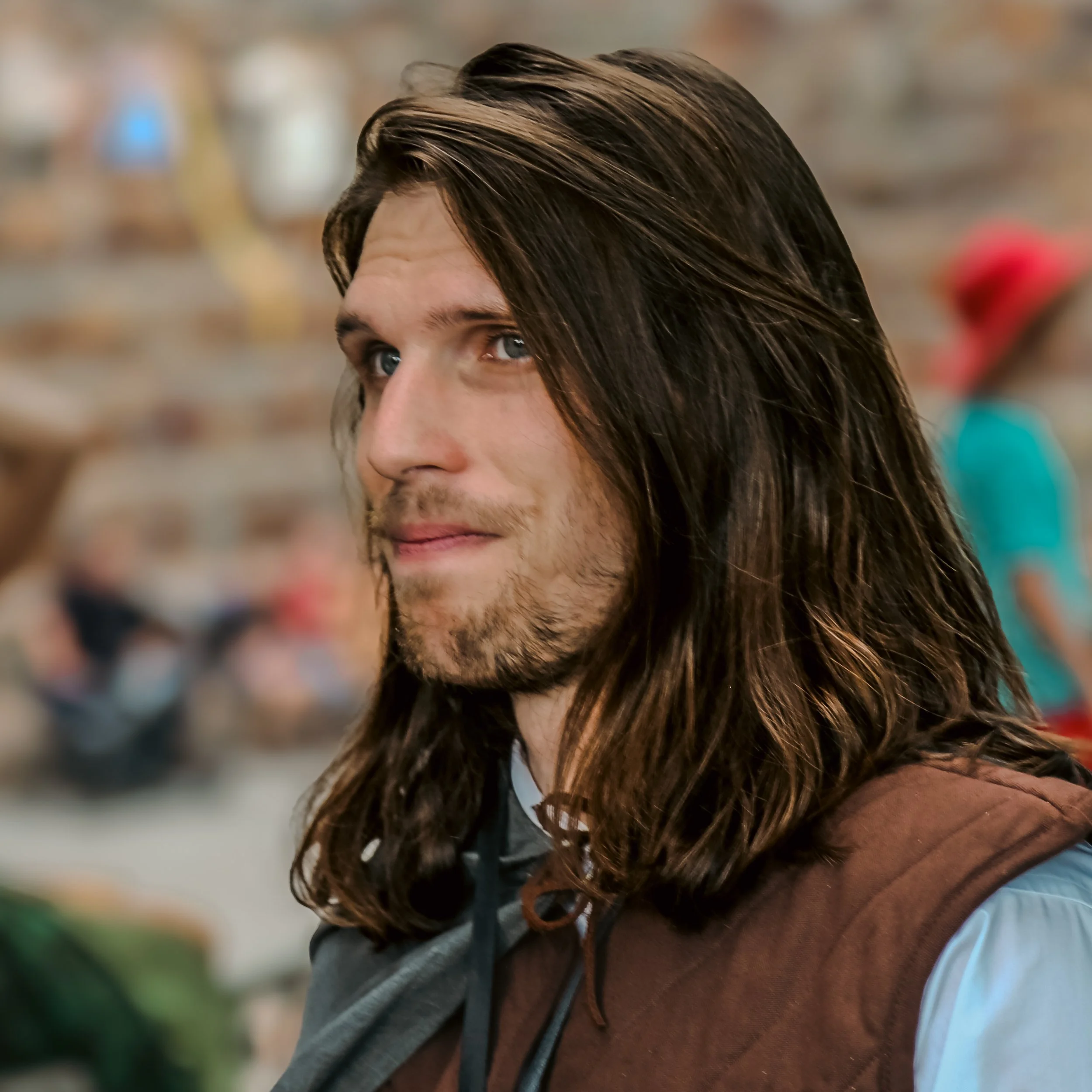 Partial view of a man with long brown hair, blue eyes, and light facial hair, wearing a gray hoodie, in an indoor shopping or store environment.