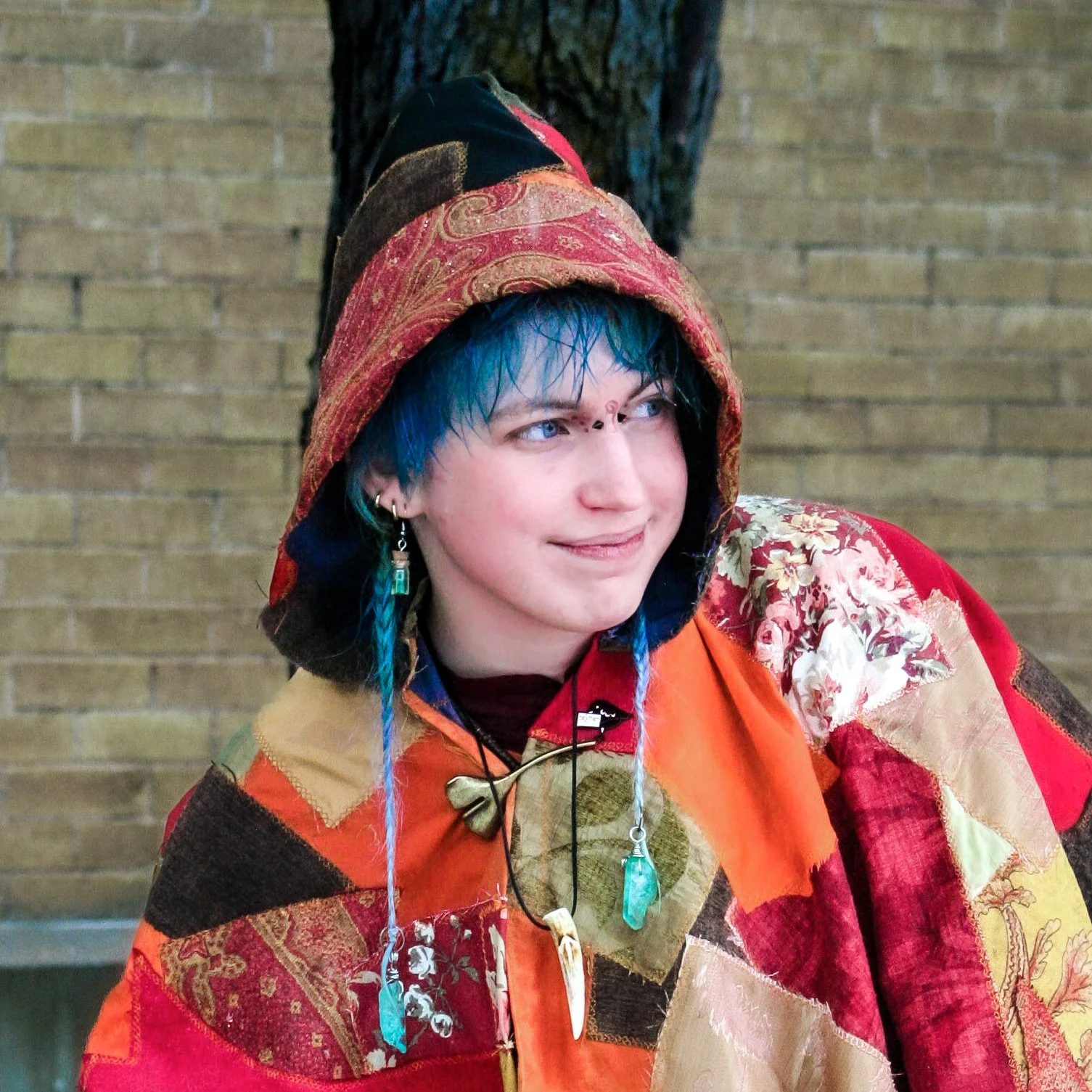 Person with dyed blue hair wearing a colorful patchwork jacket and hood, accessorized with jewelry, sitting against a brick wall.