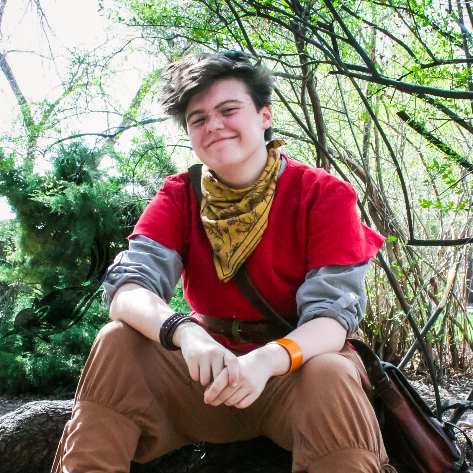 A young person with short brown hair smiling, sitting outdoors among leafy trees, wearing a red vest over a long sleeve shirt, a yellow bandana, brown pants, and black bracelets, with a backpack beside them.