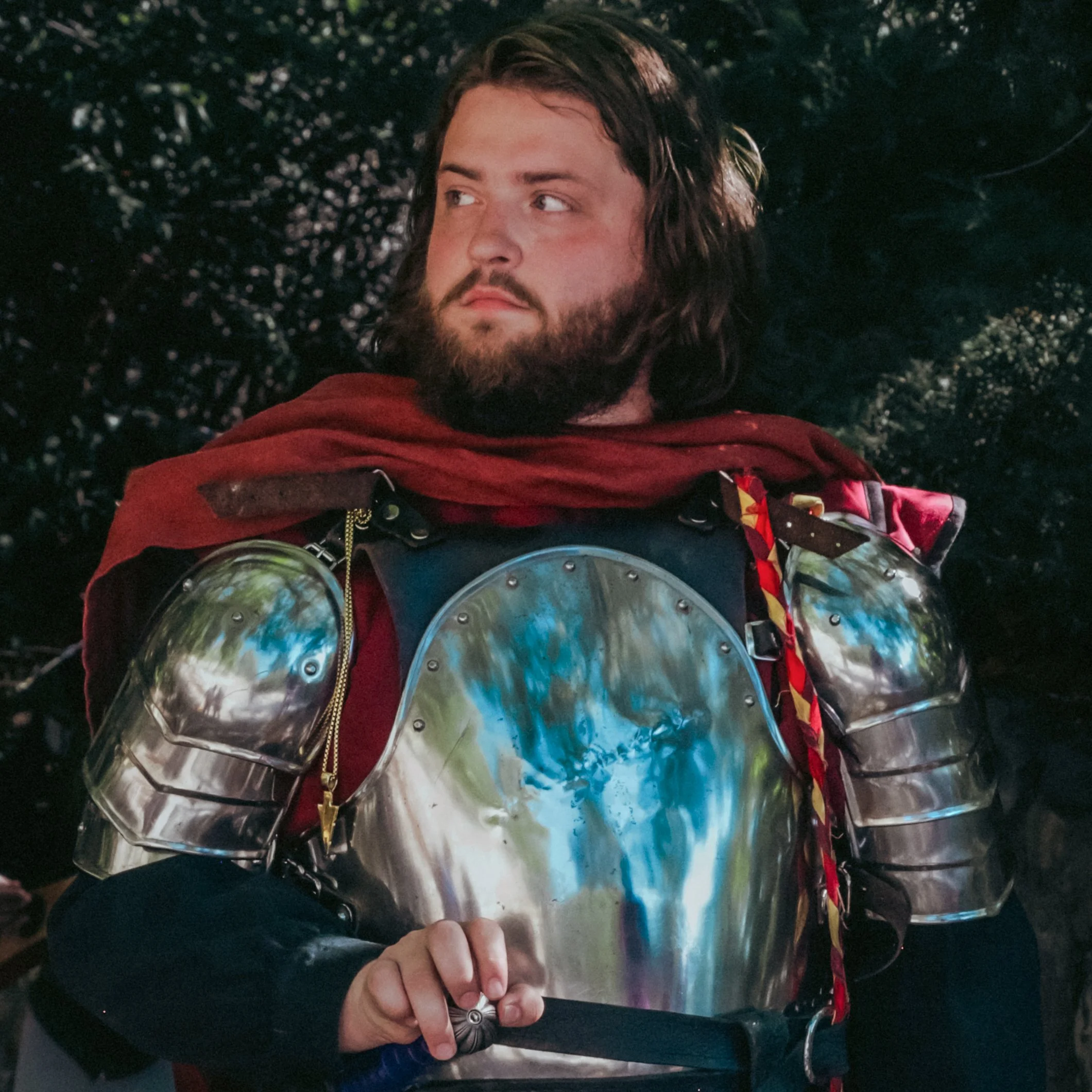A person with long brown hair and a beard is lying on a dark surface, holding a knife with a decorative handle in their right hand.