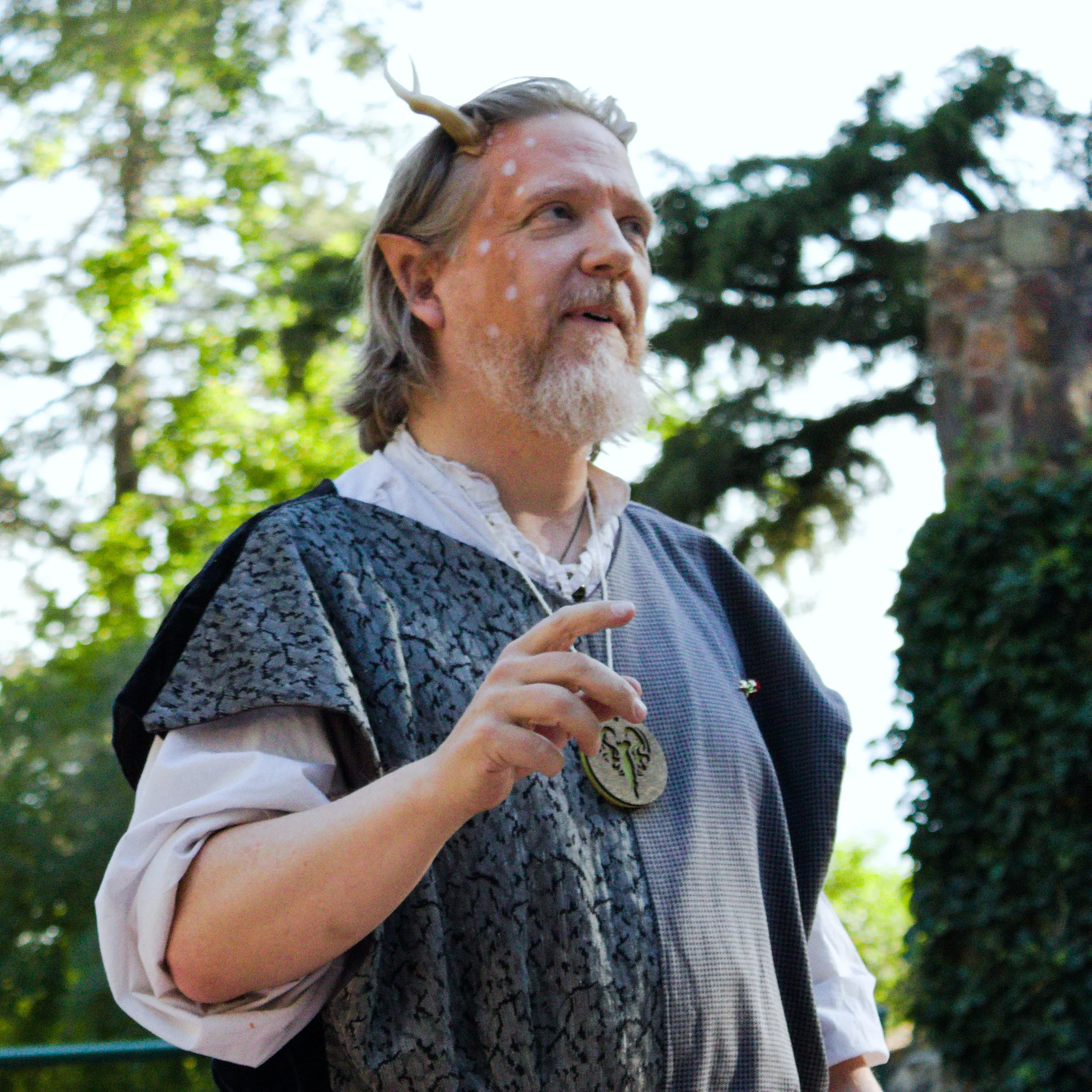 A person with pointed elf-like ears, white facial spots, and a unicorn horn headband standing outdoors among trees.