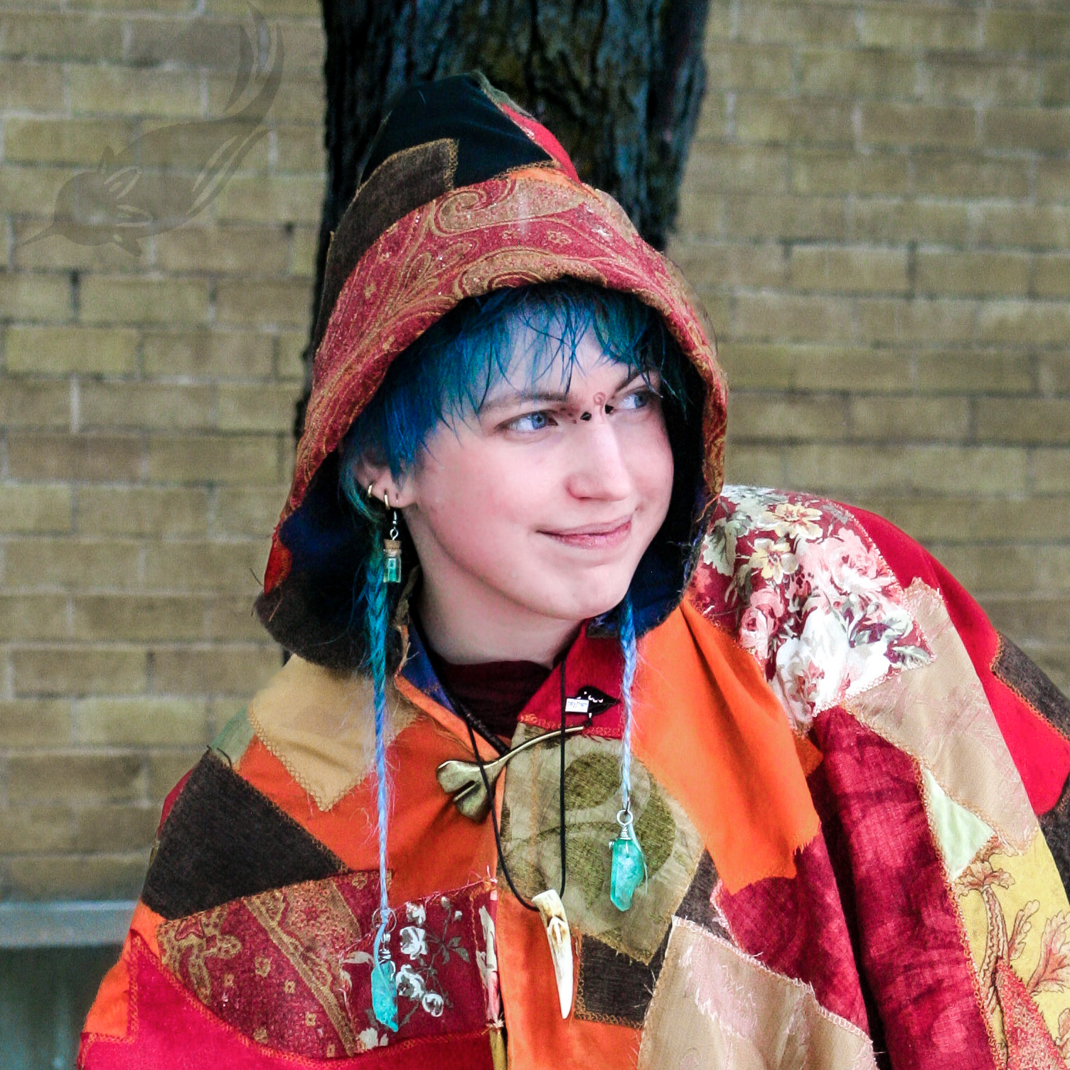 A person with blue hair wearing a colorful patchwork hoodie with a floral pattern, earrings, and a pendant necklace, standing outdoors with a brick wall and a tree in the background.