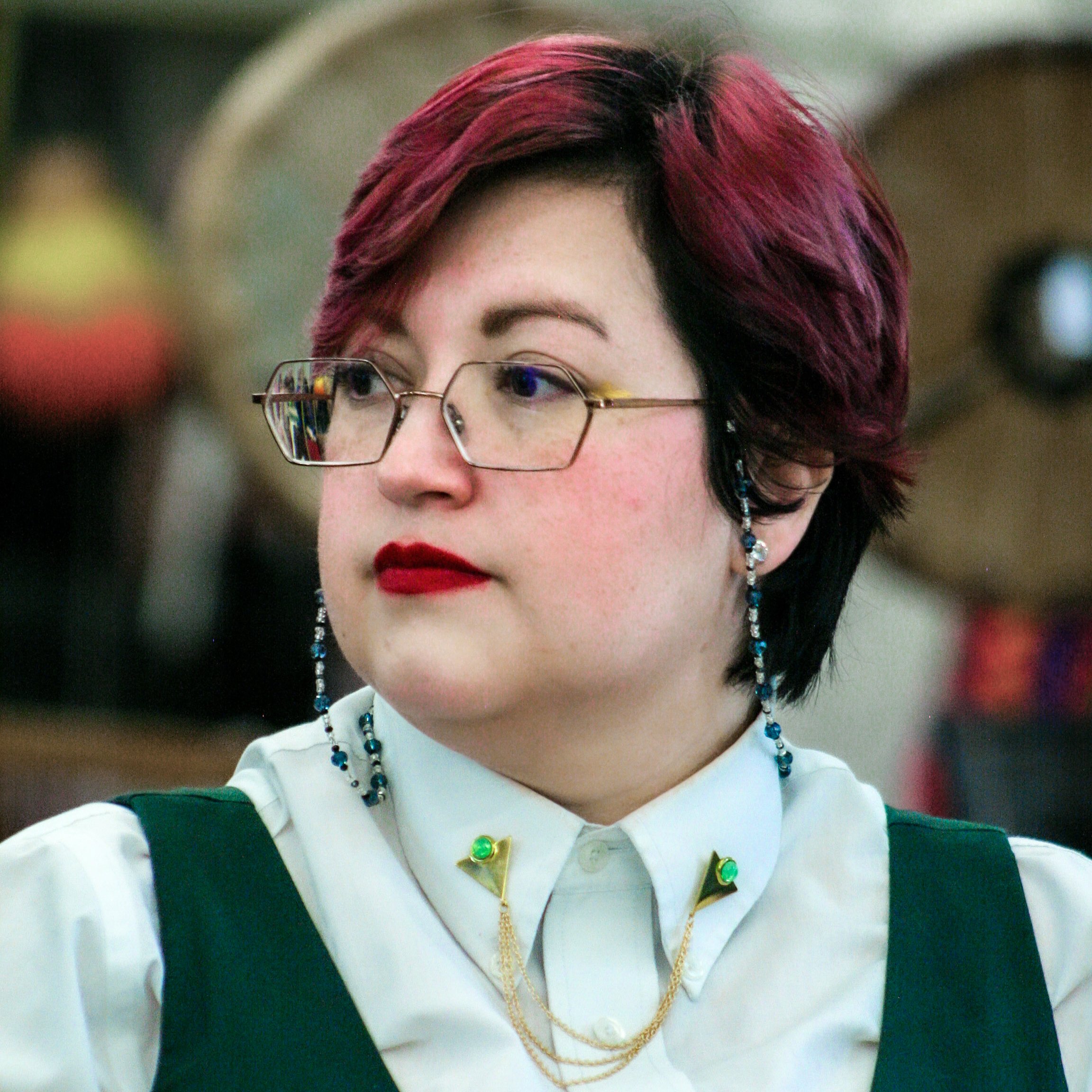 A woman with short, multicolored hair wearing glasses, a white uniform, and jewelry.