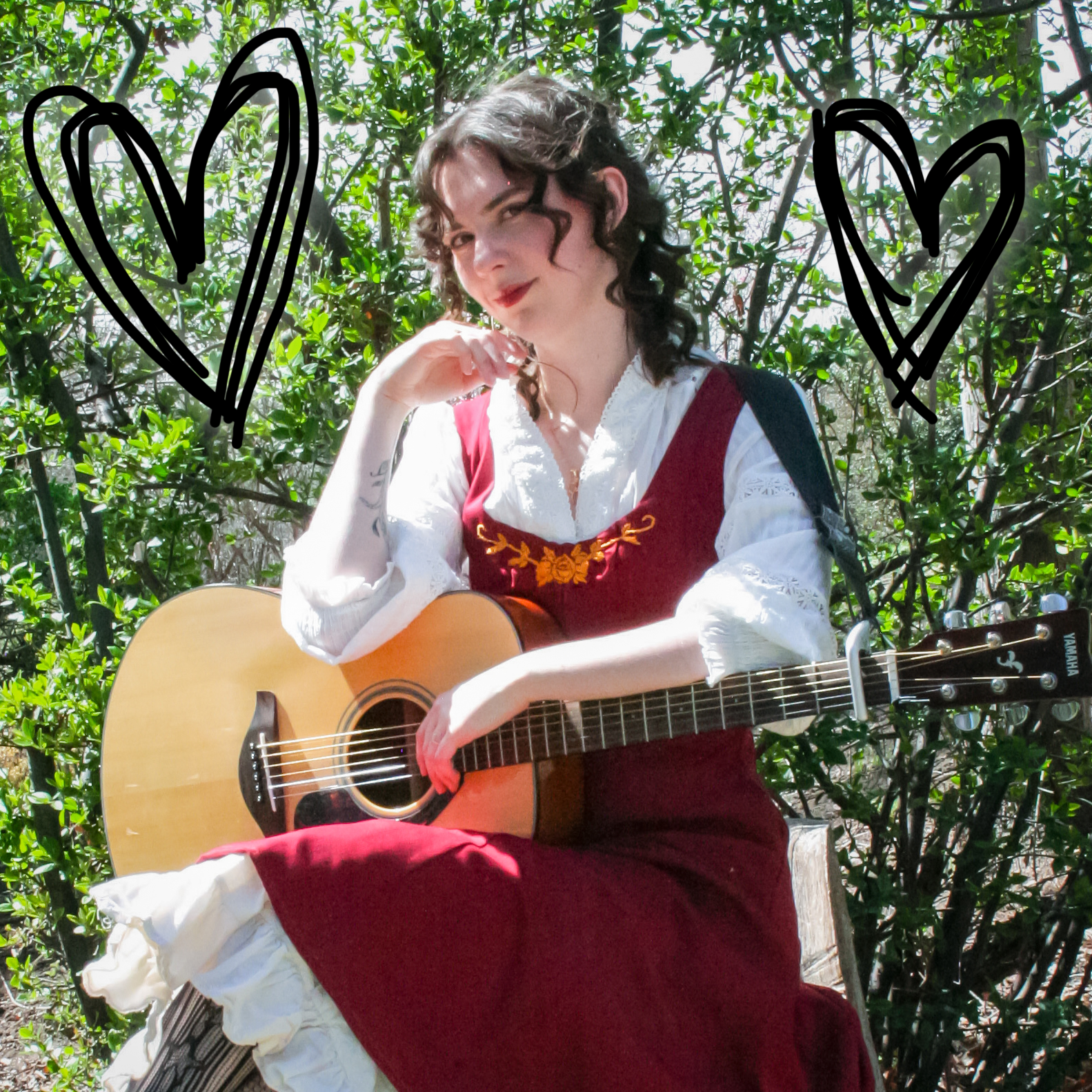 A woman with curly brown hair, wearing a white blouse and a red dress with floral embroidery, holds an acoustic guitar while sitting outdoors among green foliage.