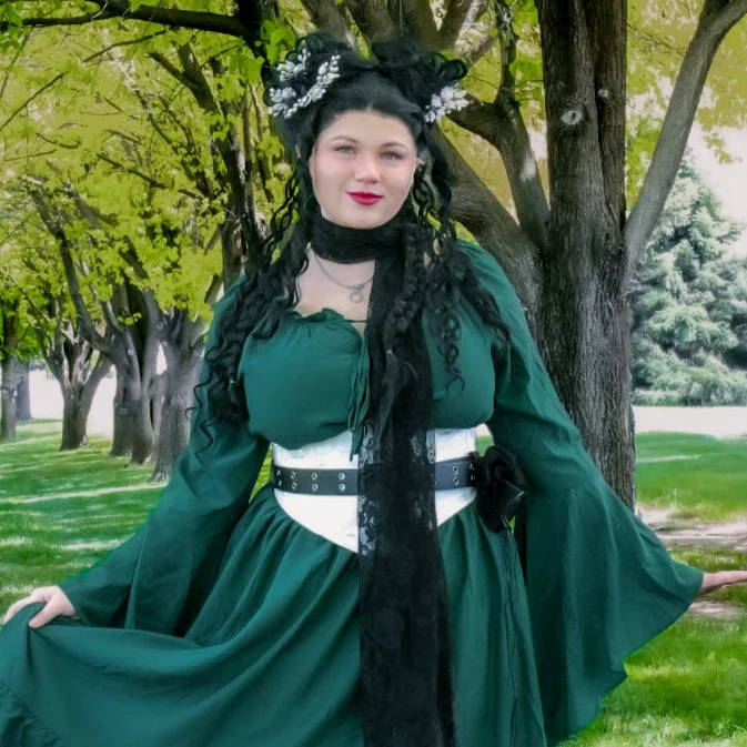 A woman dressed in a green gown with long, flowing sleeves standing outdoors near trees with green leaves.