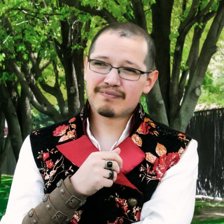 A man with glasses, a goatee, and a shaved head standing outdoors in front of green trees, wearing a black vest with red and gold floral embroidery over a white shirt, holding a red collar, and looking at the camera with a slight smile.
