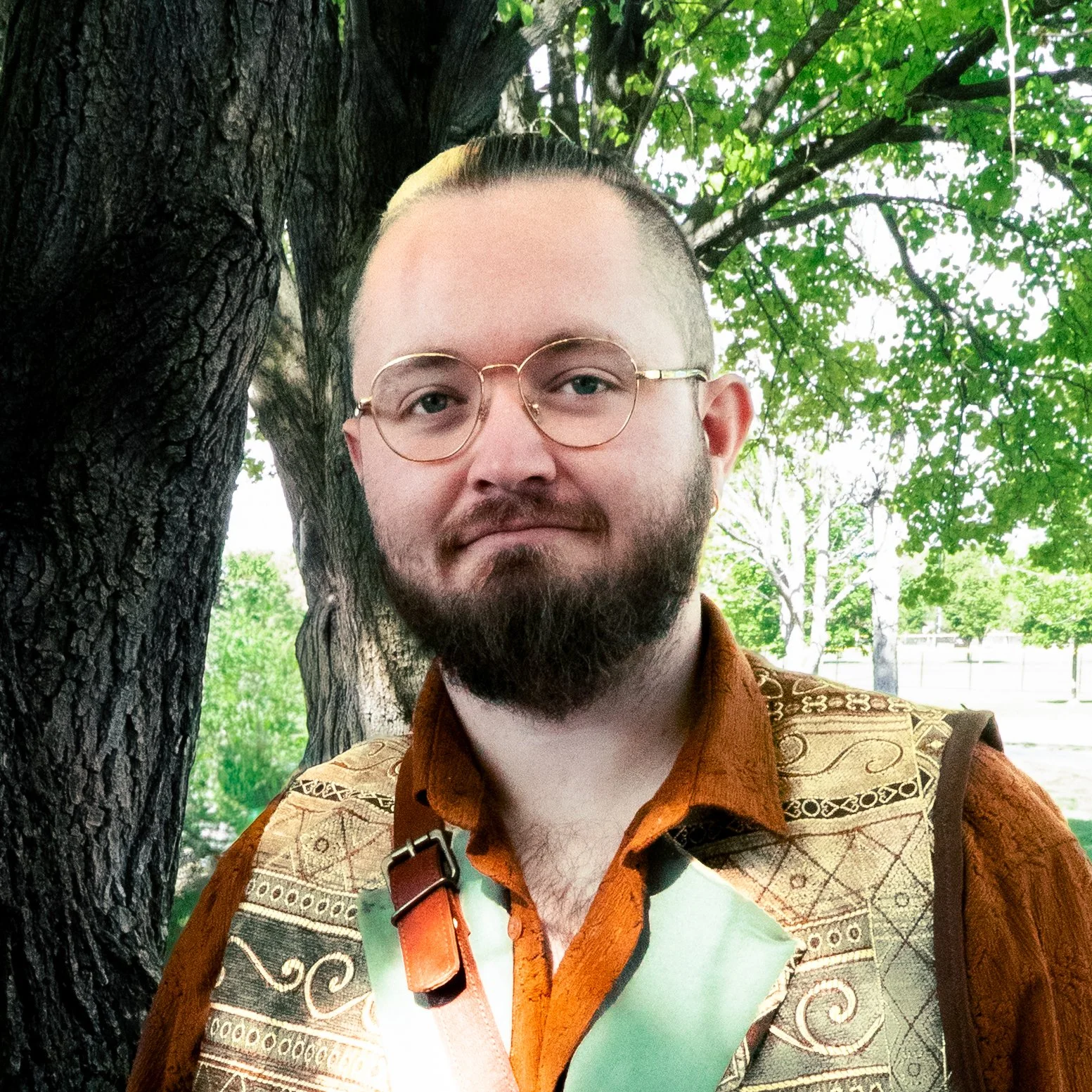 A man with glasses and a beard standing outdoors next to a large tree with green leaves, wearing a brown patterned jacket over a shirt, with a shoulder strap visible.