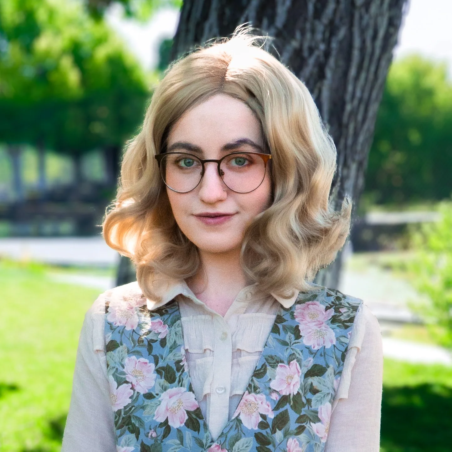 A woman with blonde, curly hair and glasses standing outdoors near a tree with a park in the background.
