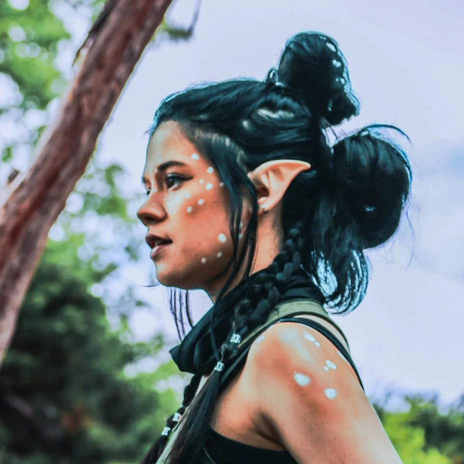 A person dressed as an elf with braided black hair, pointed ears, and face paint with white dots, standing outdoors near a tree.