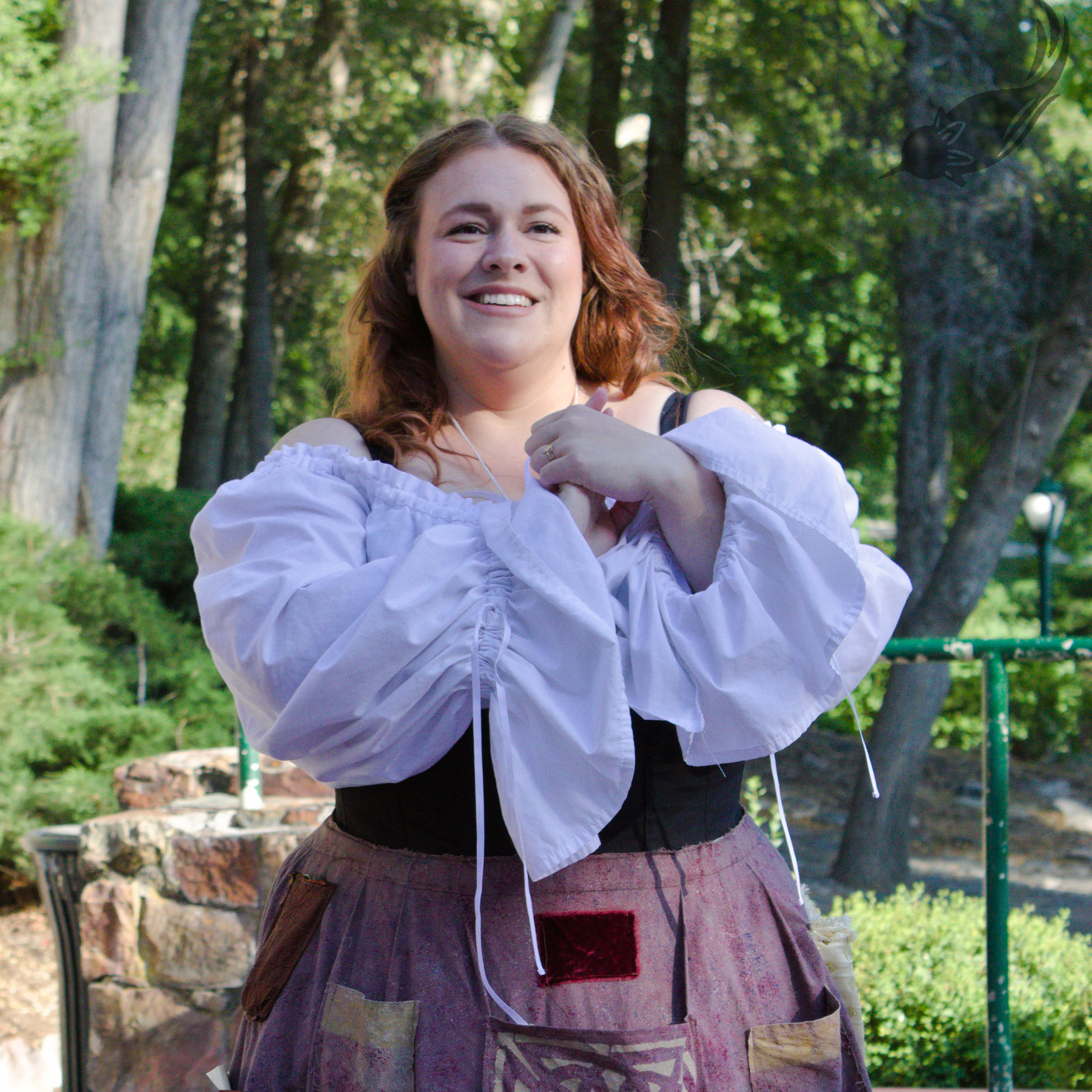 A woman with red hair smiling outdoors in a park, wearing a white off-shoulder top and an apron, holding her hands together in front of her chest.