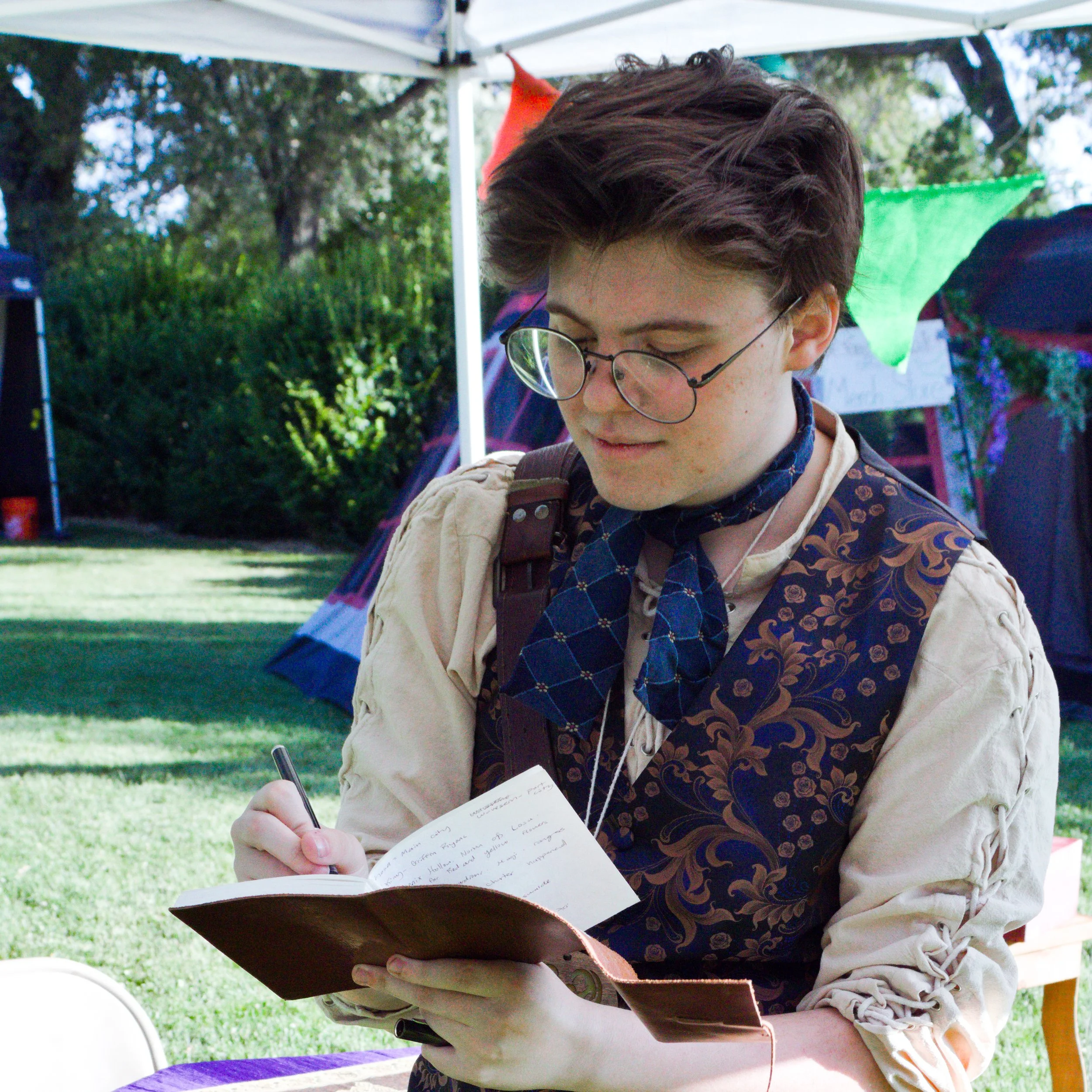 A person with short brown hair, glasses, wearing a cream-colored jacket and a patterned blue scarf, is writing on a notepad while outdoors under a white canopy. The background shows trees and colorful fabric banners.