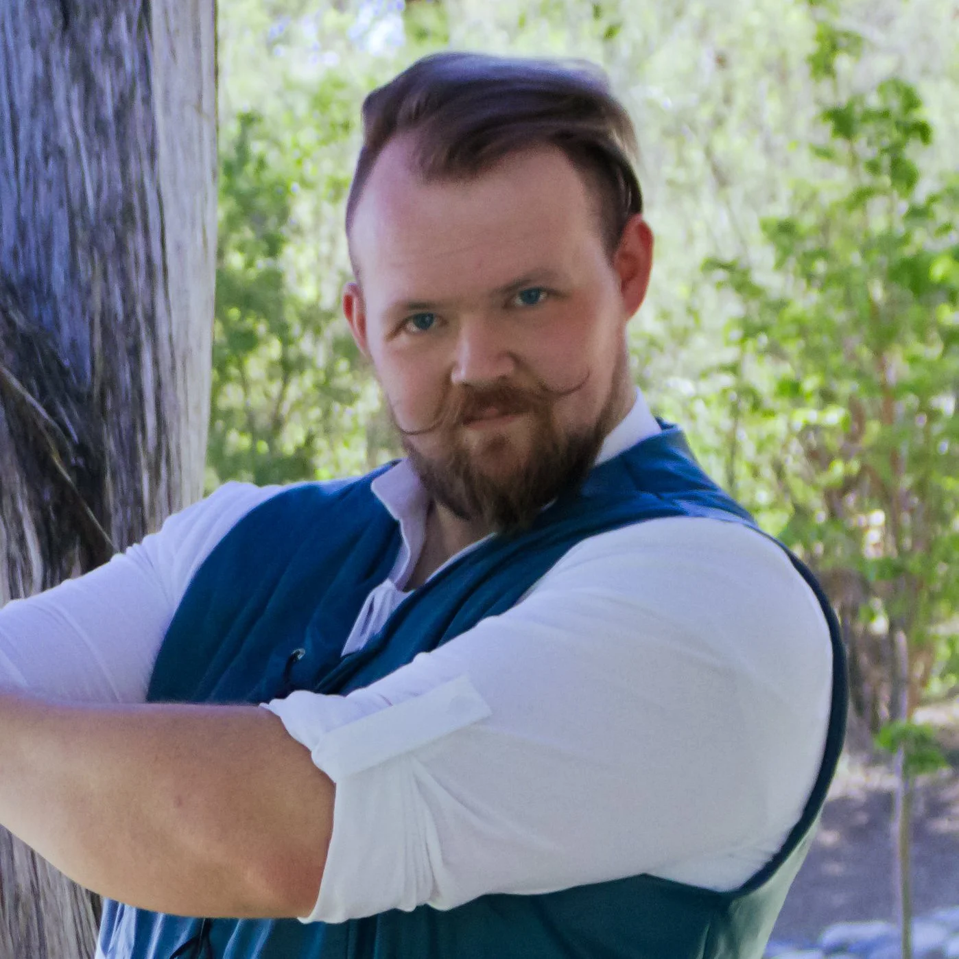 A man with styled hair, a beard, and a mustache, wearing a blue vest over a white shirt, outdoors near a tree with green foliage in the background.