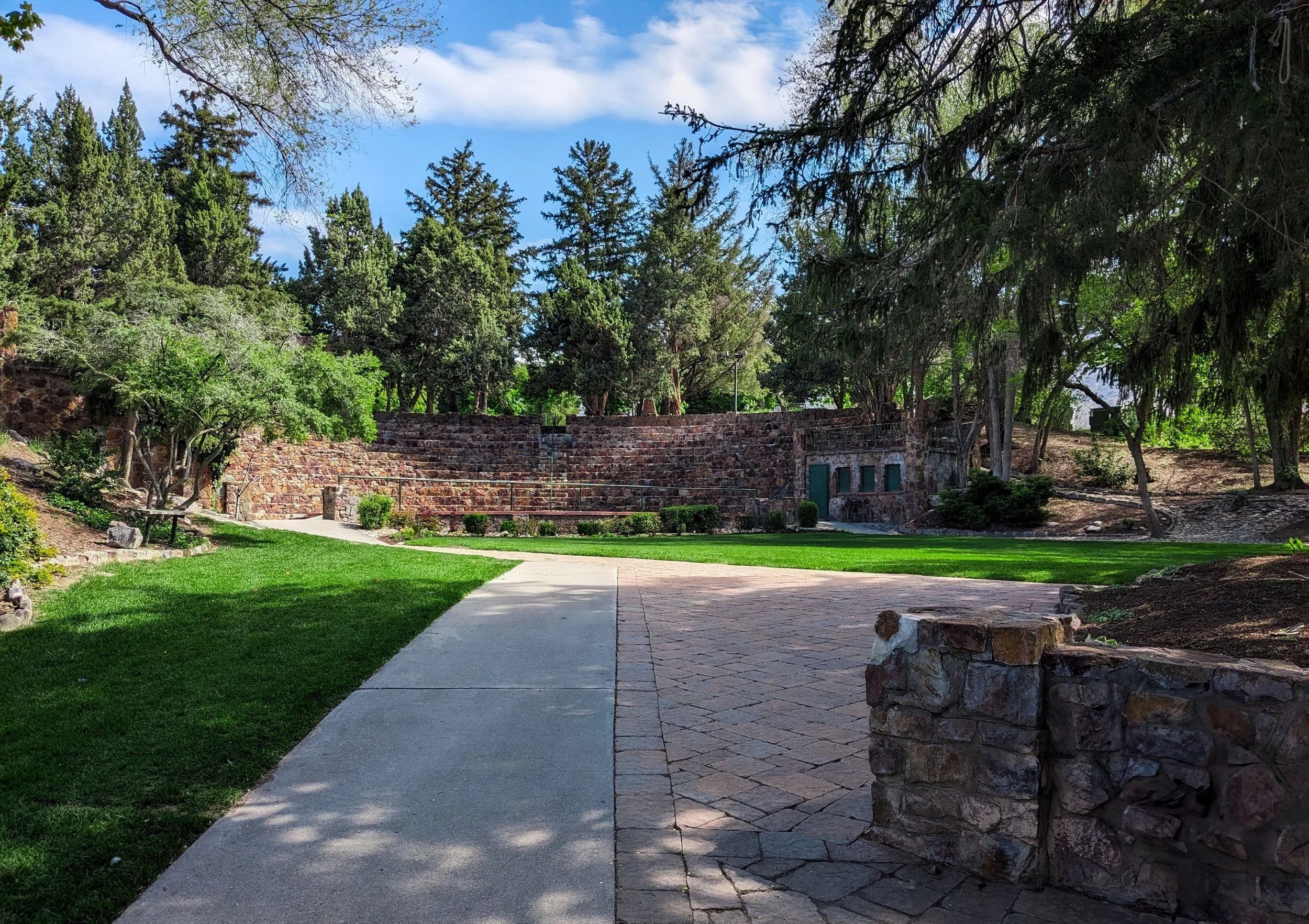a park with a paved and stone path, green grass, trees, and a stone wall with windows, under a partly cloudy sky.
