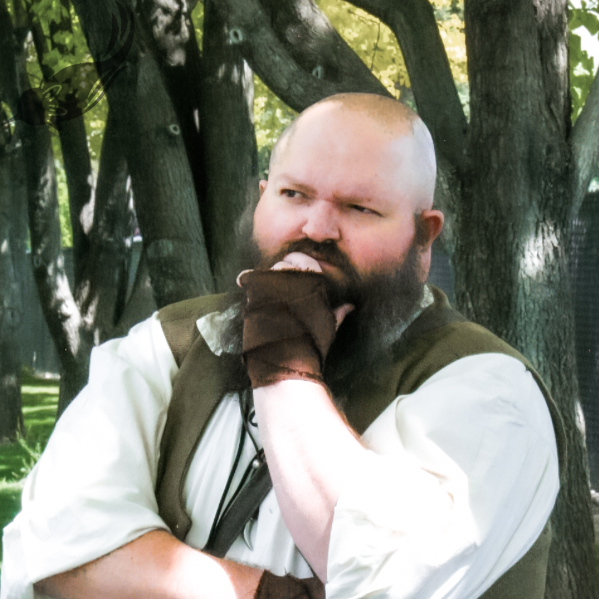 A man with a beard and shaved head wearing a medieval or fantasy-style outfit and gloves, standing outdoors with trees in the background, pretending to think or bite his glove.
