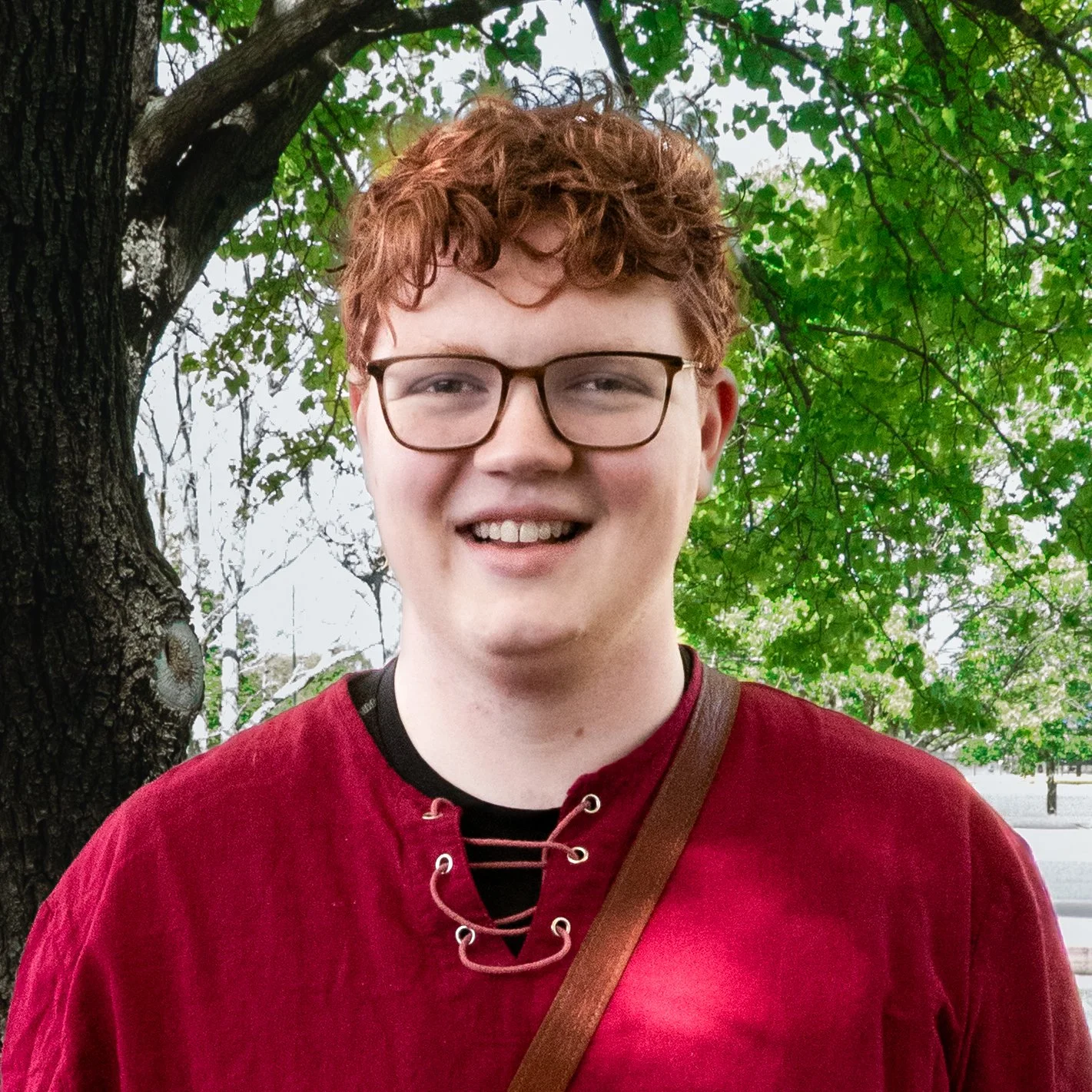 Young man with curly red hair and glasses smiling outdoors, wearing a red shirt, standing in front of a tree with green leaves.