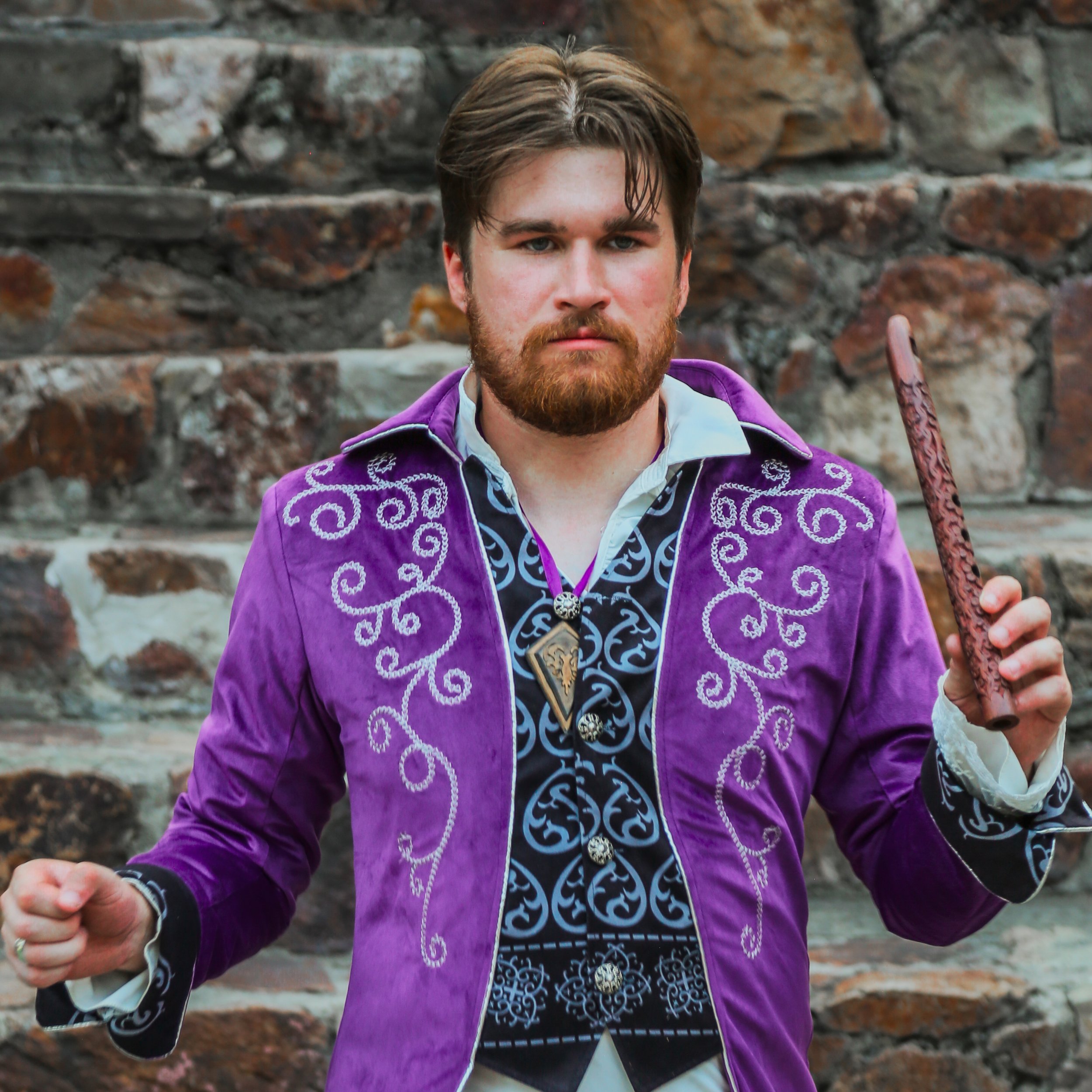 A man with brown hair and a beard is standing in front of a stone wall. He is wearing a purple embroidered jacket over a patterned shirt and holding a wooden stick.